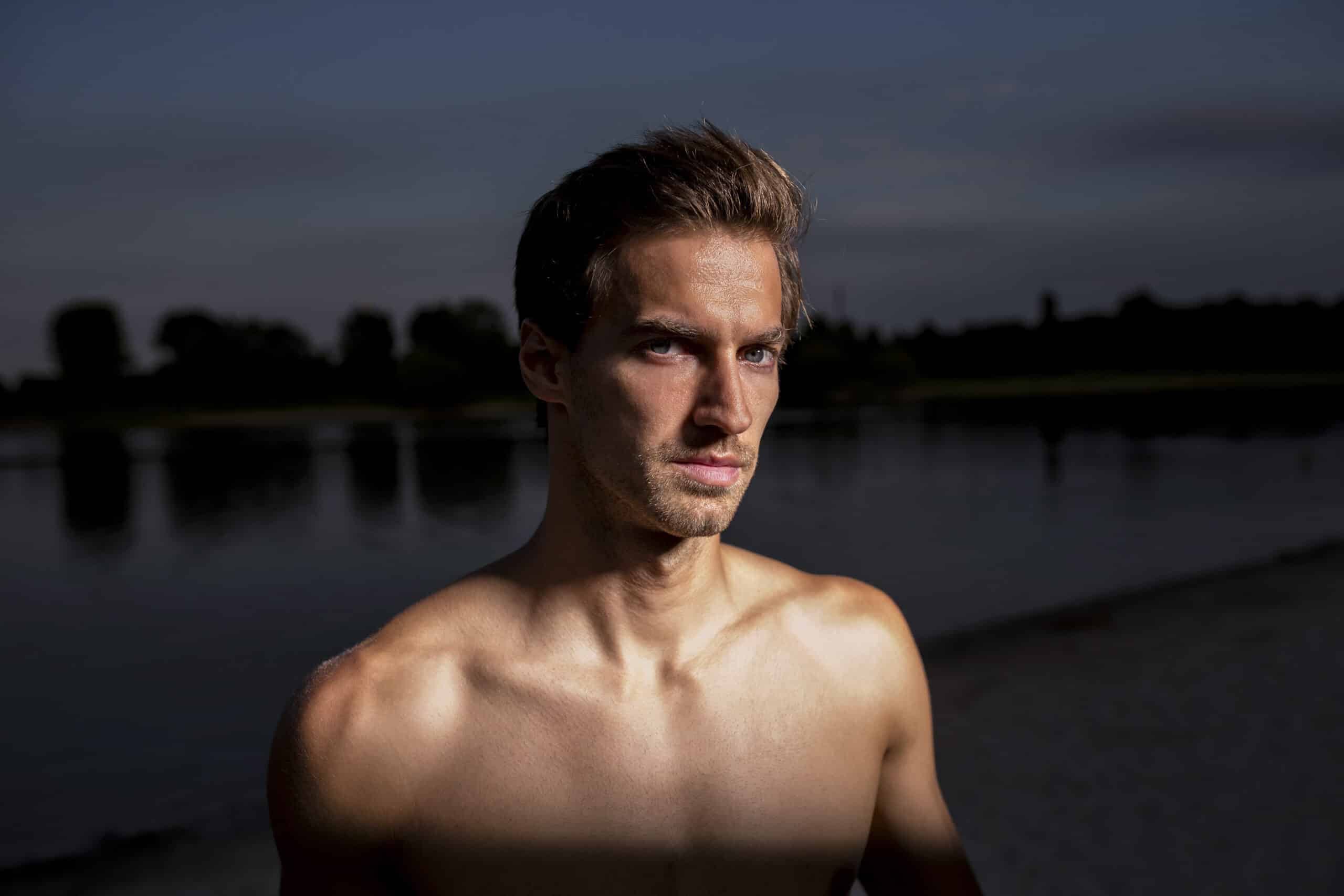 Ein Mann ohne Hemd und mit kurzen Haaren steht unter einem dunklen Himmel am Rand eines Gewässers. Der Hintergrund ist schwach beleuchtet, Bäume und eine Uferlinie sind kaum zu erkennen. Das Gesicht des Mannes ist beleuchtet, was seinen ernsten Gesichtsausdruck hervorhebt. © Fotografie Tomas Rodriguez