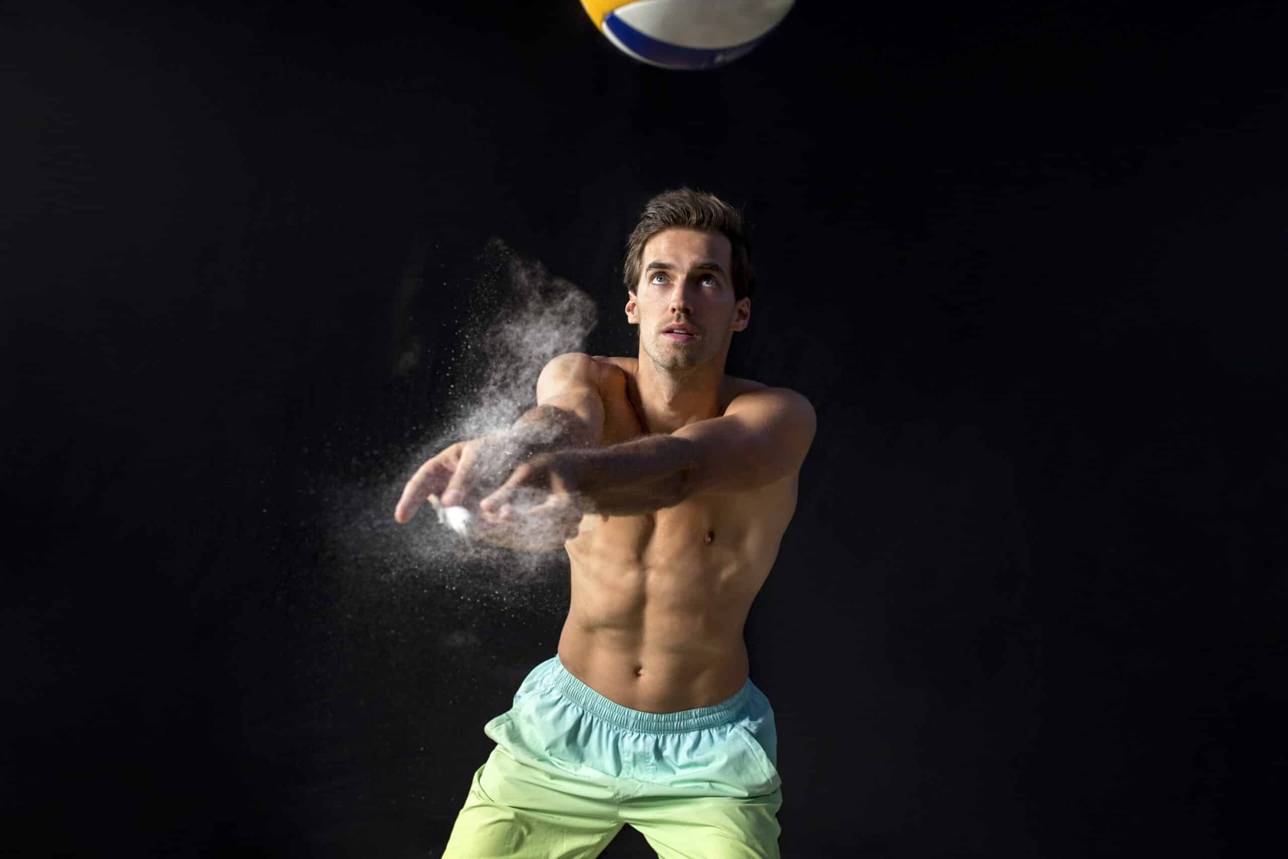 Ein muskulöser Mann ohne Hemd und in grünen Shorts ist gerade dabei, einen Volleyball zu schlagen. Er hat die Arme ausgestreckt und einen konzentrierten Gesichtsausdruck, vor einem dunklen Hintergrund und etwas weißem Kreidepulver, das aus seinen Händen rieselt. © Fotografie Tomas Rodriguez