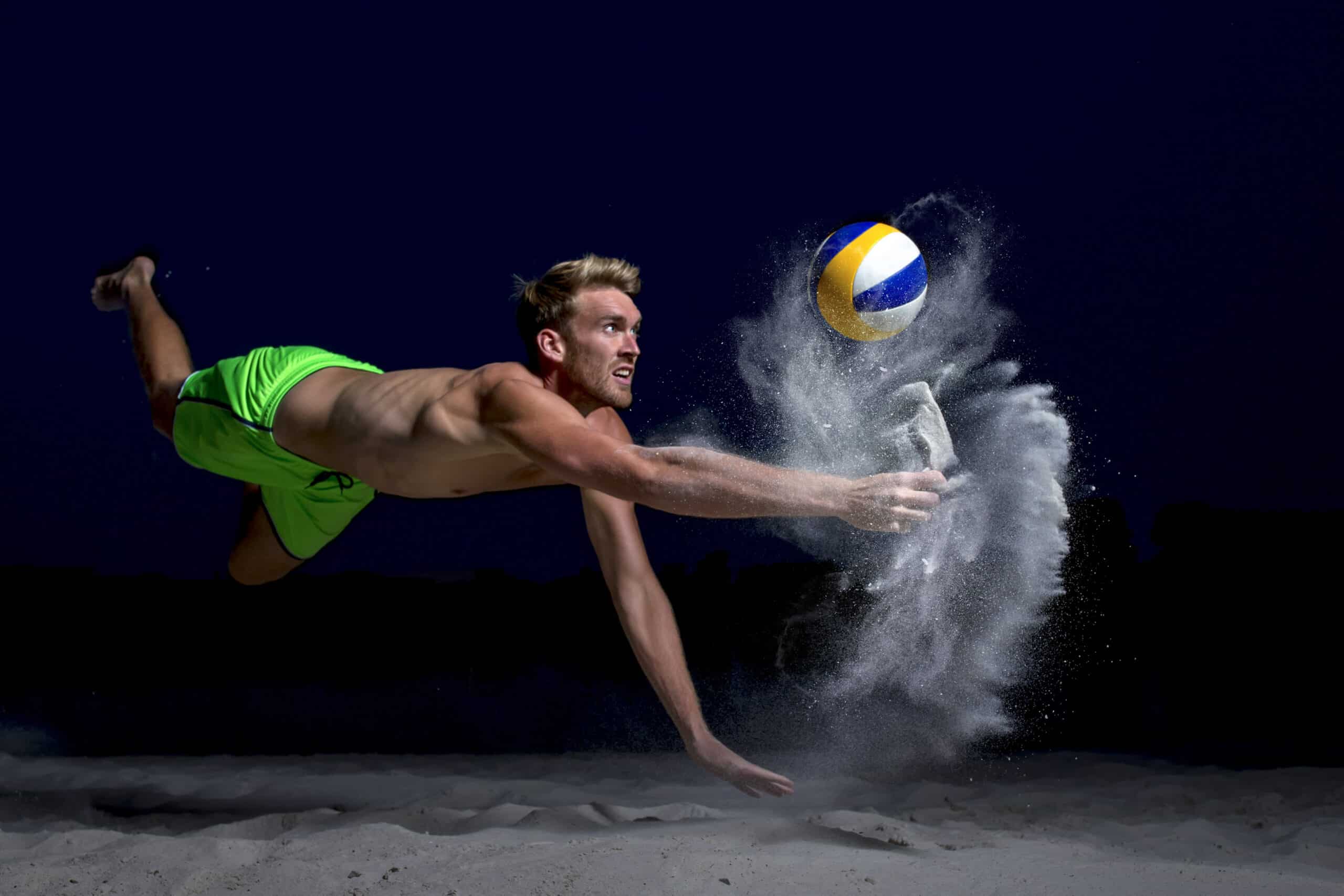 Ein Mann in leuchtend grünen Shorts stürzt sich dramatisch in die Luft, um einen Volleyball an einem Sandstrand bei Nacht zu treffen. Das Bild fängt den Moment ein, in dem er den Ball berührt, wobei Sand sichtbar um seine Hand herum verteilt wird. © Fotografie Tomas Rodriguez