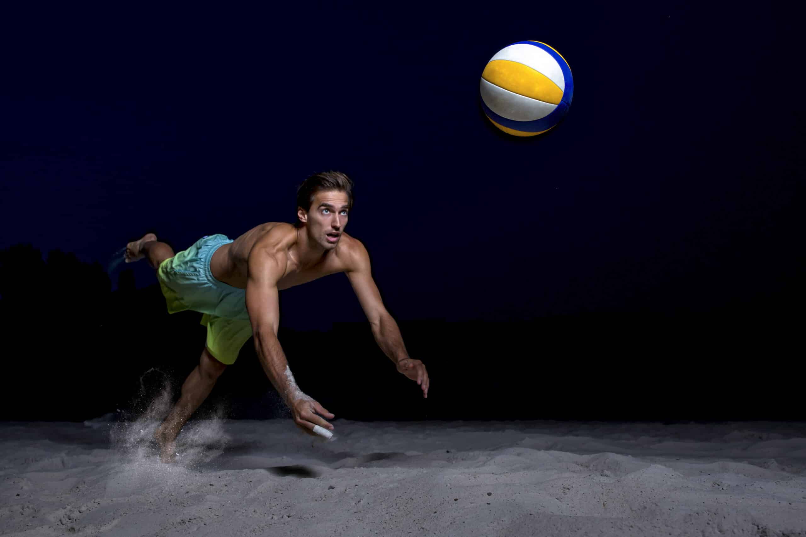 Ein Mann ohne Hemd und in grünen Shorts hechtet auf einen Sandboden und greift mit beiden Händen nach einem gelb-weißen Volleyball in der Luft, vor einem dunklen, nächtlichen Hintergrund. © Fotografie Tomas Rodriguez