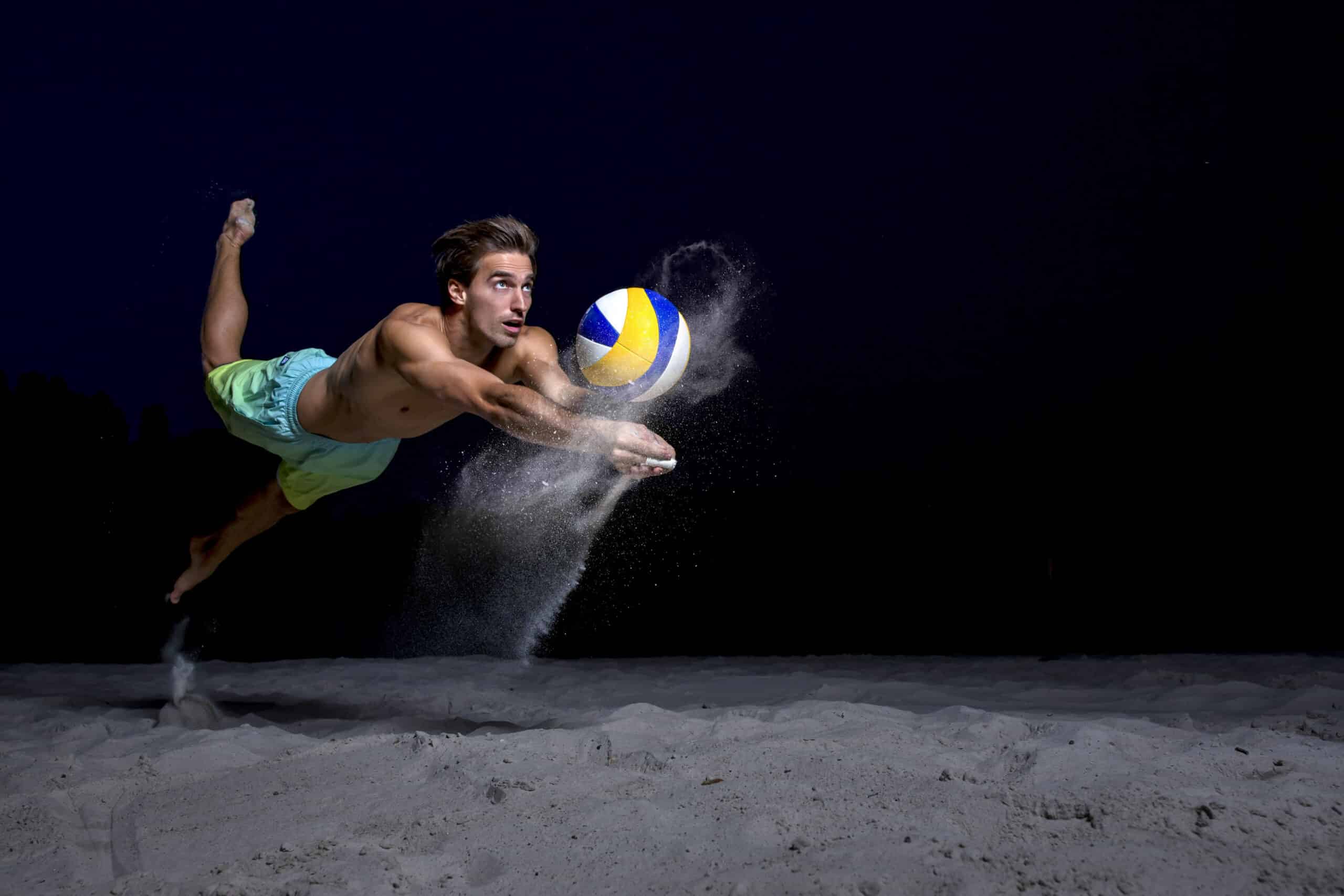 Ein Mann ohne Hemd in grünen Shorts taucht in die Luft, die Augen auf einen Volleyball gerichtet, die Arme ausgestreckt, um den Ball zu schlagen. Durch seine Bewegung wird Sand aufgewirbelt. Der Hintergrund ist dunkel, was die dramatische Action der Beachvolleyball-Szene hervorhebt. © Fotografie Tomas Rodriguez