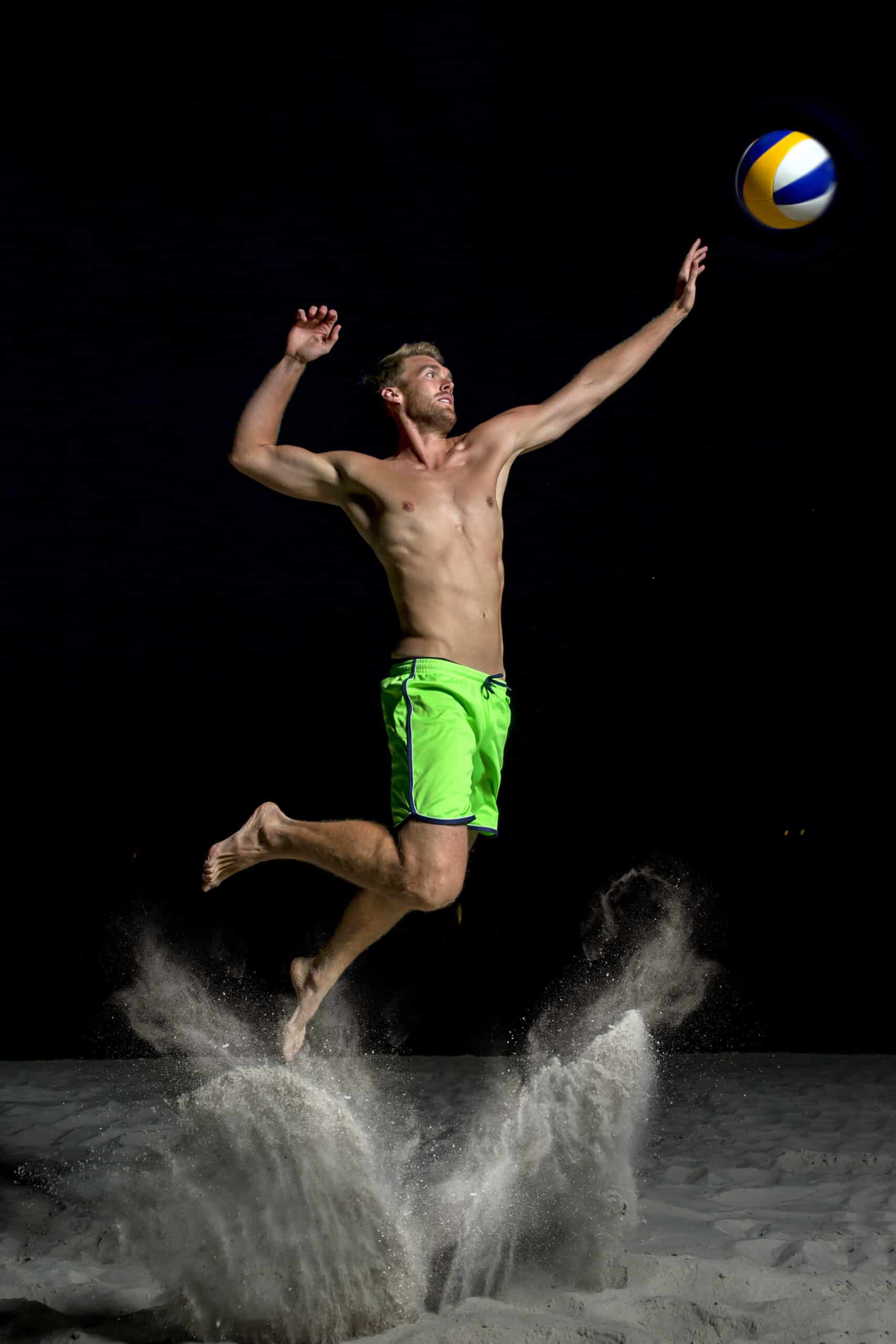 Ein Mann ohne Hemd und in leuchtend grünen Shorts wird nachts beim Beachvolleyballspielen in der Luft fotografiert. Er springt hoch, einen Arm ausgestreckt, und versucht, den Ball zu schlagen. Unter ihm wirbelt Sand auf, was die Intensität seines Sprungs unterstreicht. © Fotografie Tomas Rodriguez
