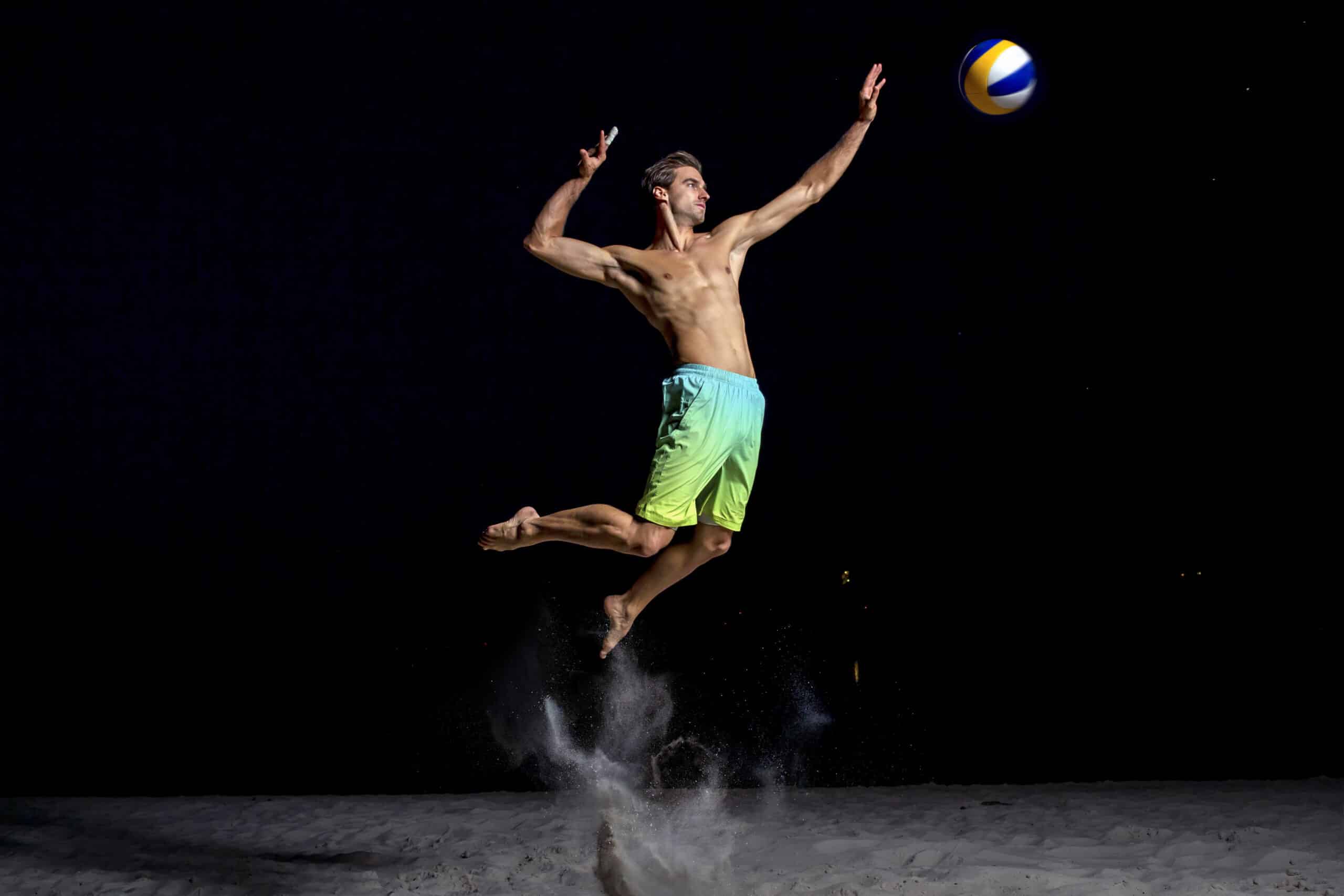 Ein Mann ohne Hemd in bunten Shorts springt nachts in die Luft, um einen Volleyball auf einem Sandboden zu schlagen. Der Ball befindet sich in der Luft und Sand wird unter ihm aufgewirbelt. Der Hintergrund ist dunkel, was die dynamische Bewegung hervorhebt. © Fotografie Tomas Rodriguez