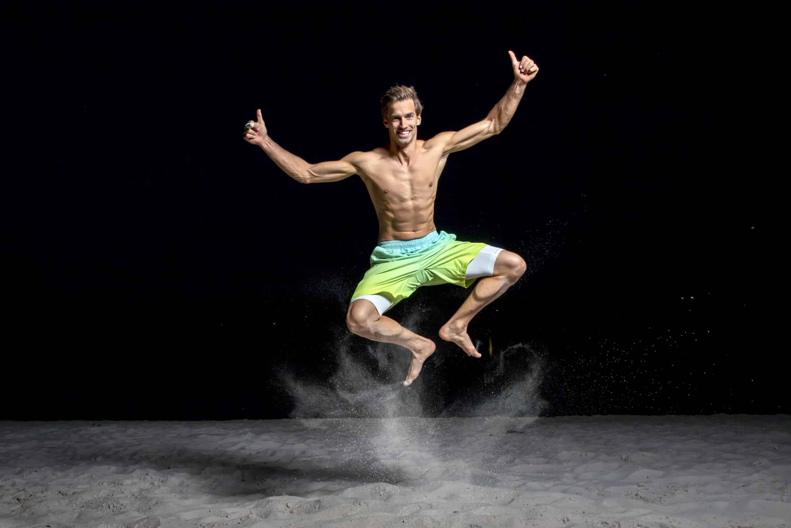 Ein Mann ohne Hemd und in neongrünen Shorts wird nachts mitten im Sprung an einem Sandstrand aufgenommen. Er hat eine athletische Figur, lächelt und hat beide Daumen hoch. Sand wird um seine Füße herum aufgewirbelt, was der Szene dynamische Energie verleiht. Der Hintergrund ist völlig dunkel. © Fotografie Tomas Rodriguez