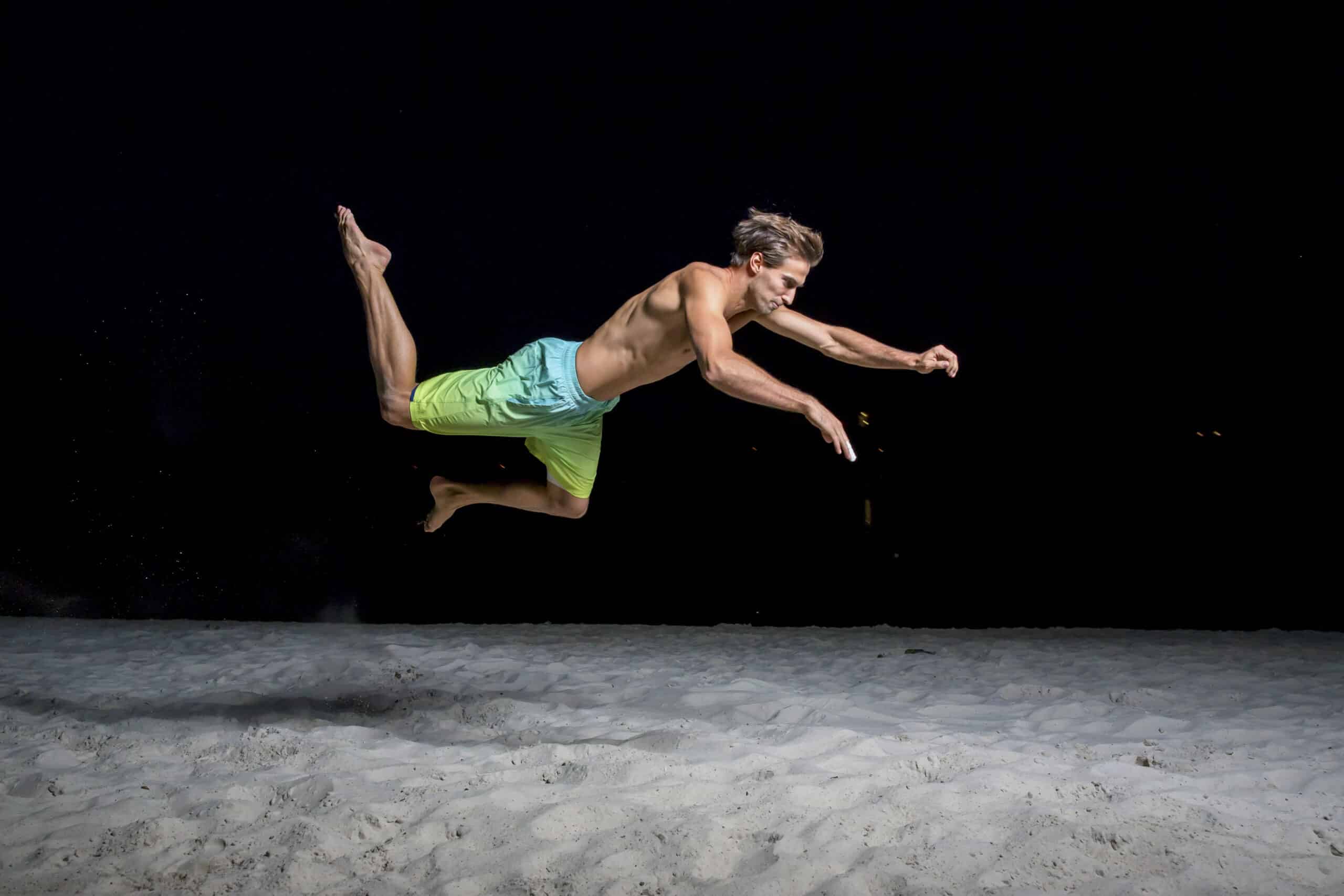 Ein Mann in neongrüner Badehose ist nachts in der Luft aufgenommen, parallel zum Boden ausgestreckt mit ausgestreckten Armen und Beinen. Er scheint über einen Sandstrand zu springen oder zu tauchen, beleuchtet vor einem dunklen Hintergrund. © Fotografie Tomas Rodriguez