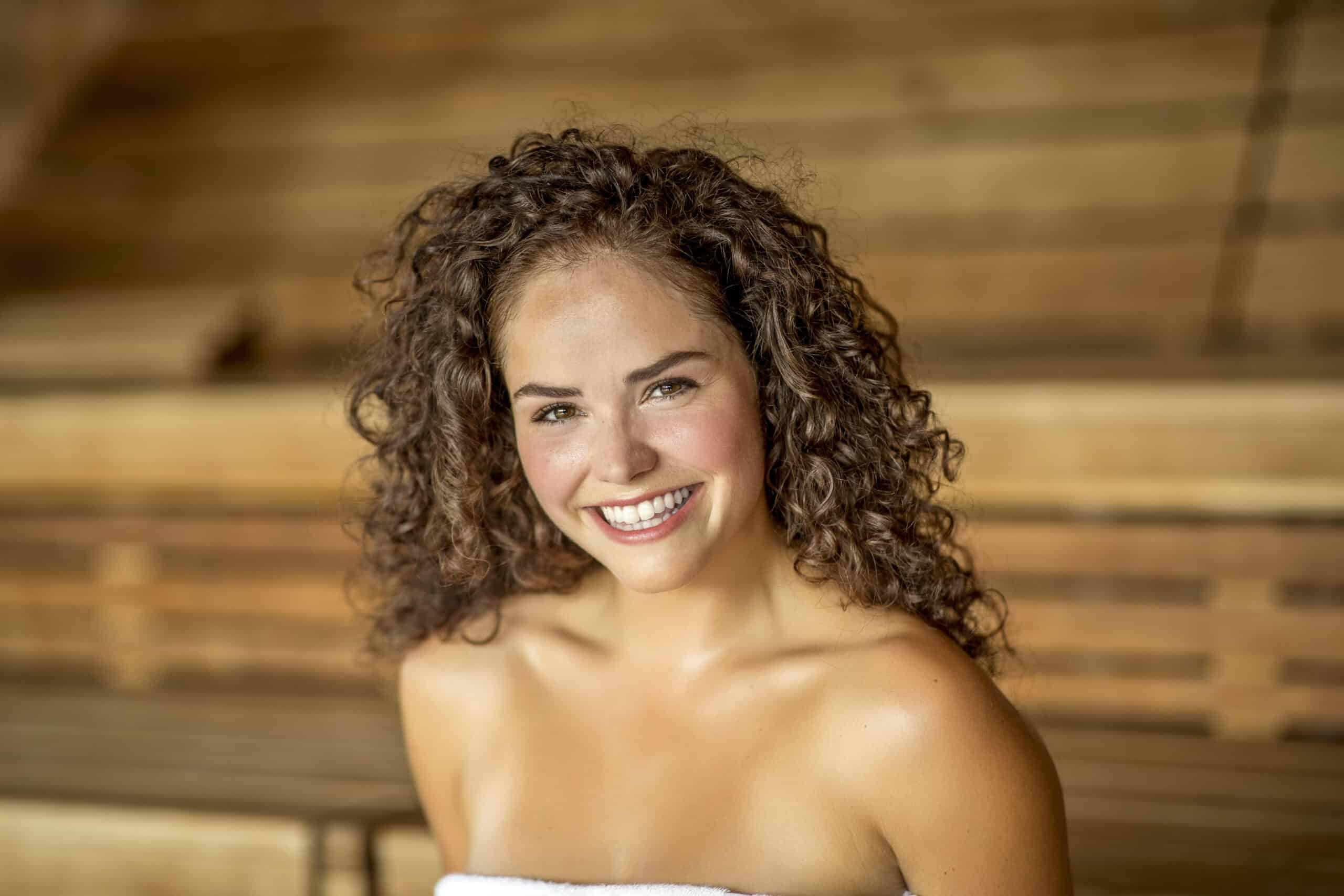 Eine Frau mit lockigem braunem Haar und strahlendem Lächeln ist in ein weißes Handtuch gehüllt. Im Hintergrund sind Holzpaneele zu sehen, die vermuten lassen, dass sie sich in einer Sauna oder einem Holzraum befindet. © Fotografie Tomas Rodriguez