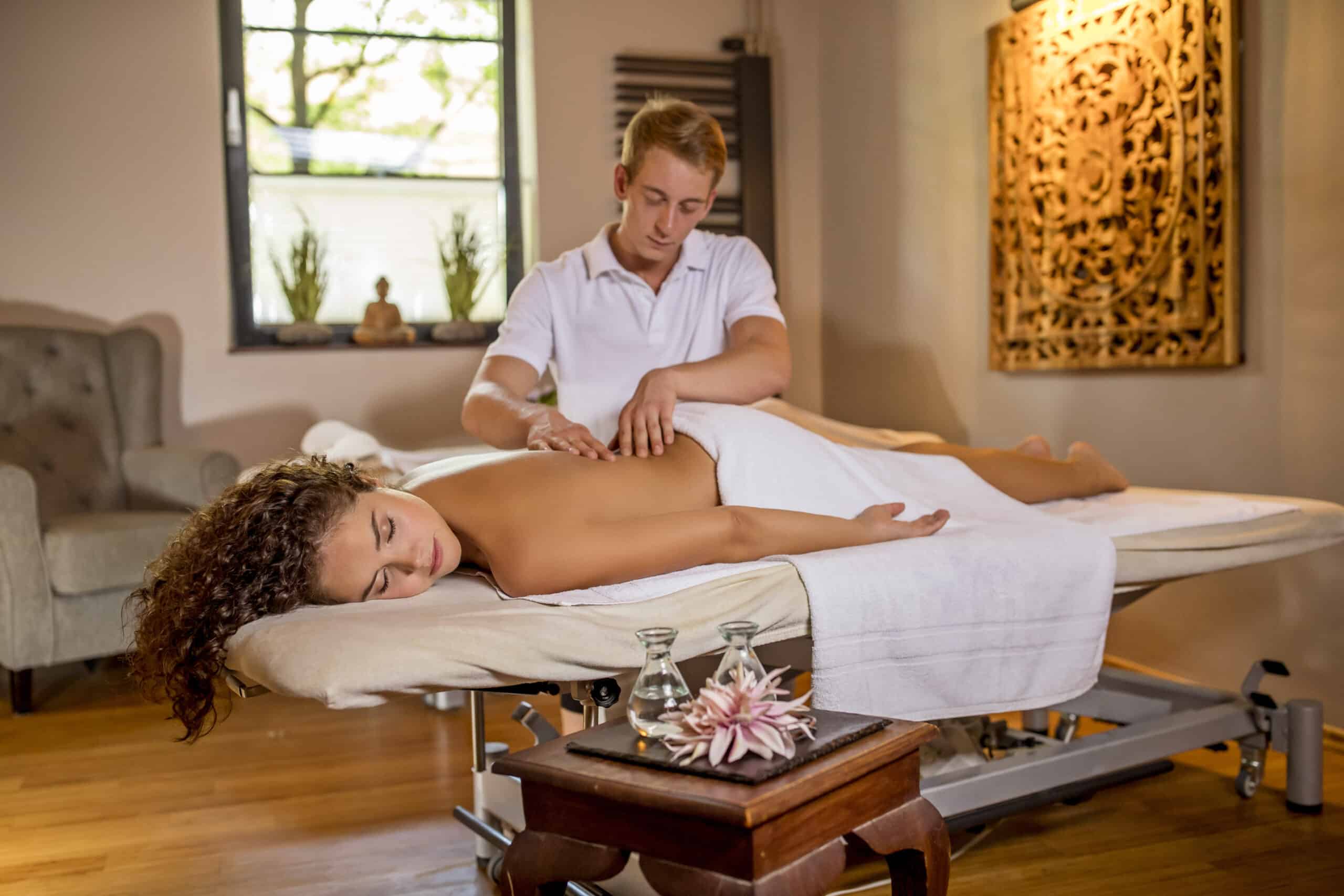 Eine Person liegt mit dem Gesicht nach unten auf einem Massagetisch, über ihren Unterkörper ist ein weißes Handtuch gehängt. Daneben behandelt ein Masseur im weißen Hemd ihren Rücken. Der Raum ist durch ein Fenster, eine geschnitzte Wanddekoration aus Holz und einen kleinen Tisch mit Massageölen sanft beleuchtet. © Fotografie Tomas Rodriguez