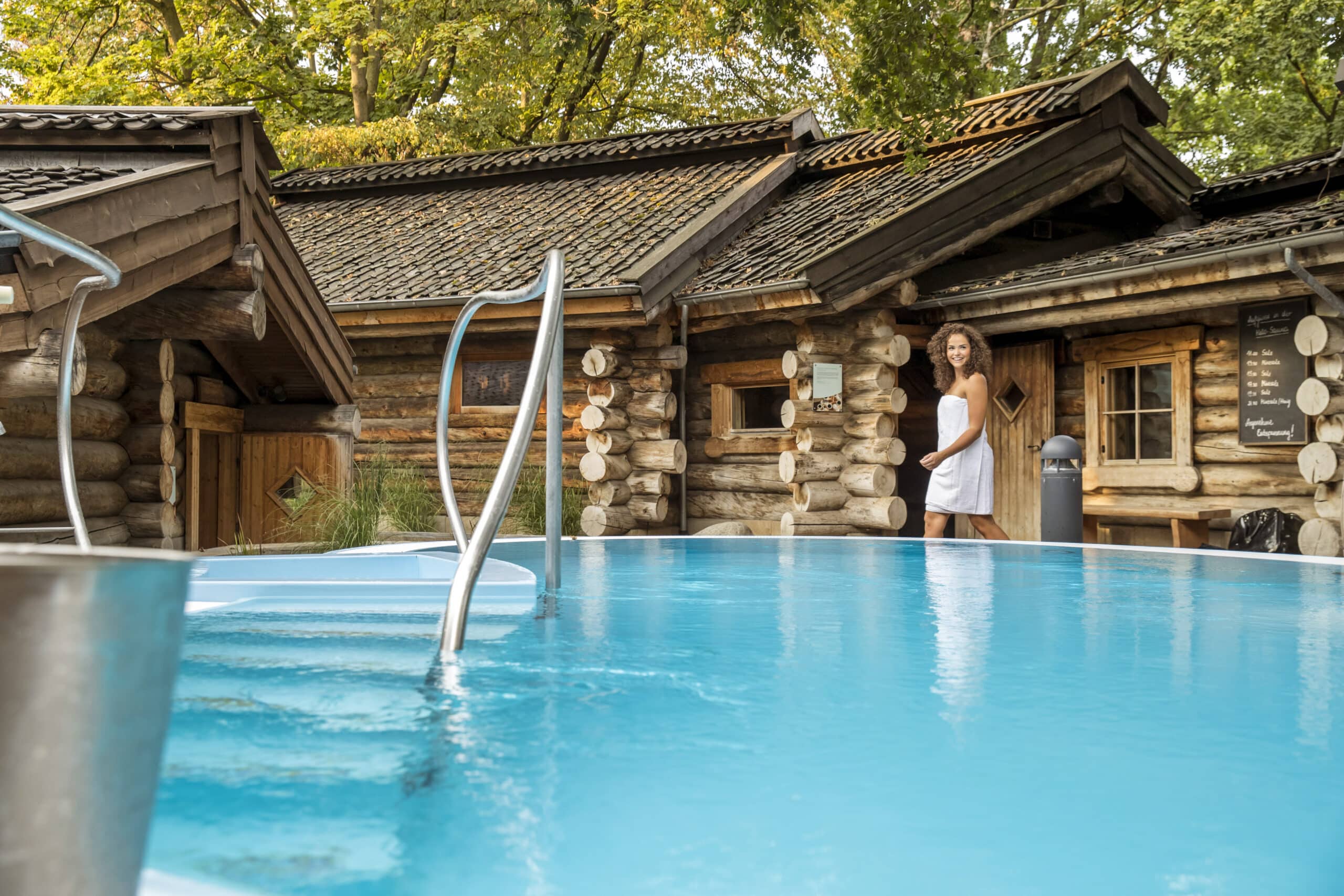 Eine in ein Handtuch gehüllte Frau steht an der Tür einer rustikalen Holzhütte. Im Vordergrund ist ein klarer, blauer Swimmingpool mit Metallgeländern zu sehen. Im Hintergrund sind Bäume mit grünem Laub zu sehen, die eine ruhige und natürliche Atmosphäre schaffen. © Fotografie Tomas Rodriguez
