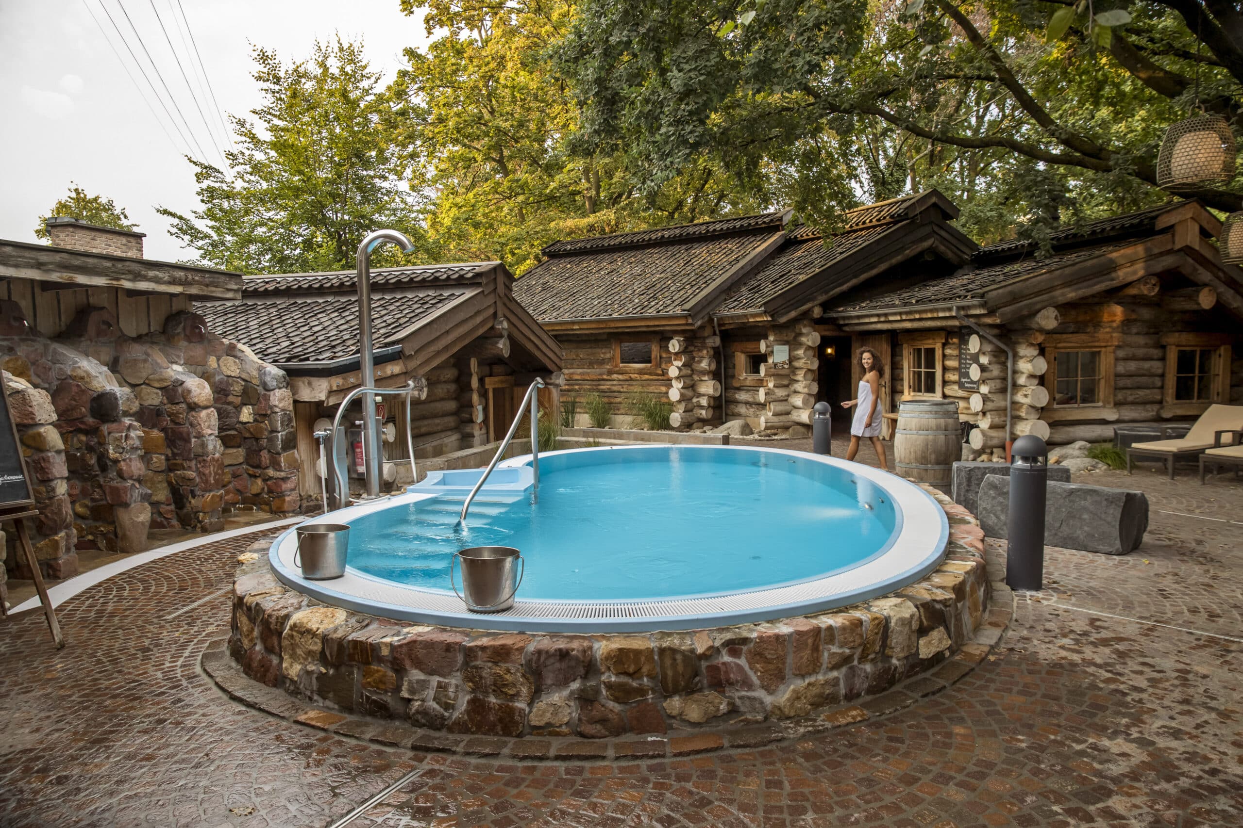 Ein ruhiges Spa im Freien verfügt über einen runden, erhöhten Pool, der von rustikalen Holzhütten umgeben ist. Der Poolbereich ist mit Kopfsteinpflaster gepflastert und im Hintergrund stehen Bäume. Am Pool sind ein Metallgeländer und ein Ausguss angebracht und eine Person steht in der Nähe und genießt die ruhige Umgebung. © Fotografie Tomas Rodriguez