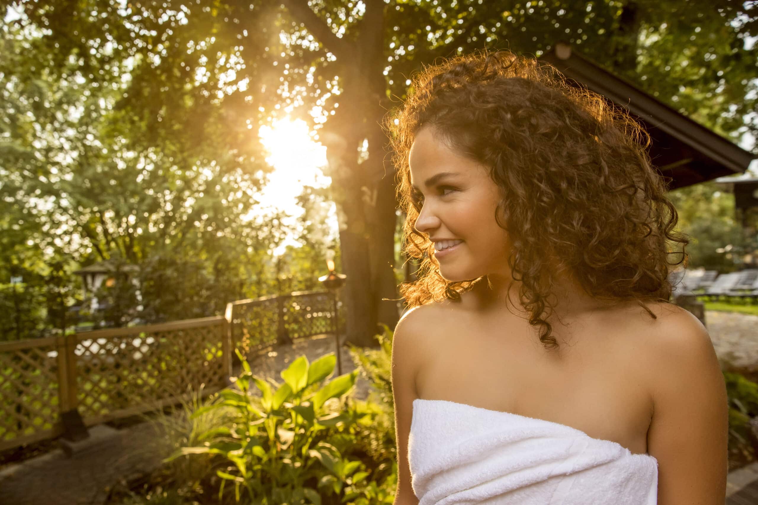 Eine Frau mit lockigem Haar und einem weißen Handtuch um sie gewickelt, schaut zur Seite und lächelt, während sie draußen in einem sonnigen Garten steht. Im Hintergrund sind üppiges Grün, ein Holzzaun und Sonnenlicht zu sehen, das durch die Bäume fällt. © Fotografie Tomas Rodriguez