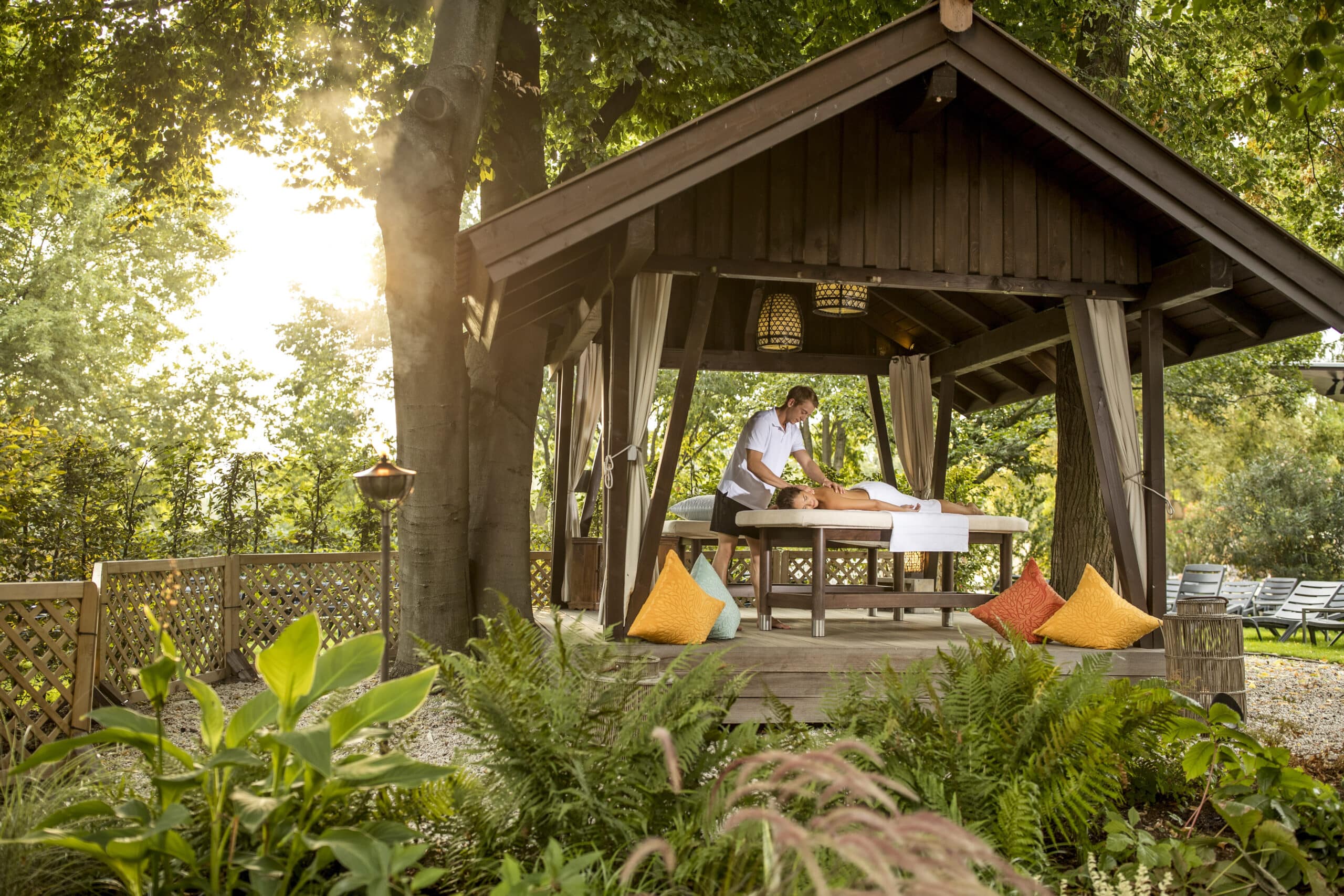 Eine ruhige Massage im Freien unter einem hölzernen Pavillon, umgeben von üppigem Grün. Der Pavillon ist mit hellen Vorhängen und farbenfrohen Kissen geschmückt und sorgt für eine friedliche Atmosphäre. Sonnenlicht fällt durch die Bäume und verleiht der Szene einen Hauch von Wärme. © Fotografie Tomas Rodriguez