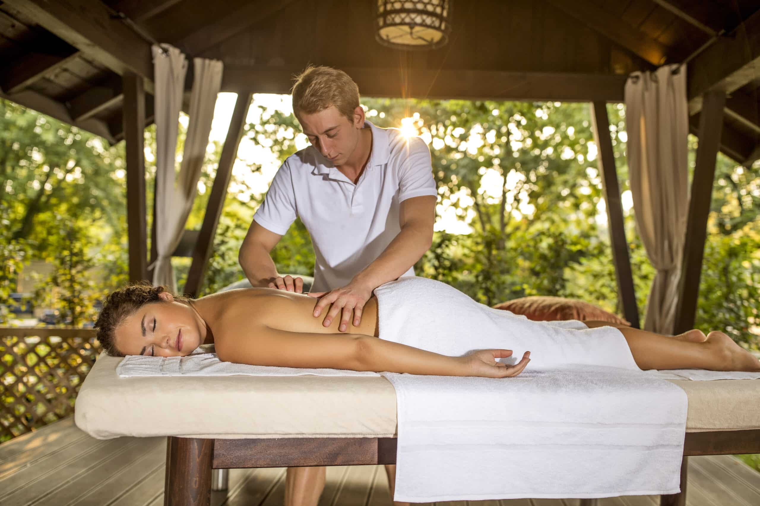 Ein Masseur massiert einer Person den Rücken, die auf einem mit einem weißen Handtuch bedeckten Massagetisch liegt. Sie befindet sich in einem Pavillon im Freien mit Vorhängen und Grün im Hintergrund. Das Sonnenlicht fällt durch die Bäume und schafft eine ruhige Atmosphäre. © Fotografie Tomas Rodriguez
