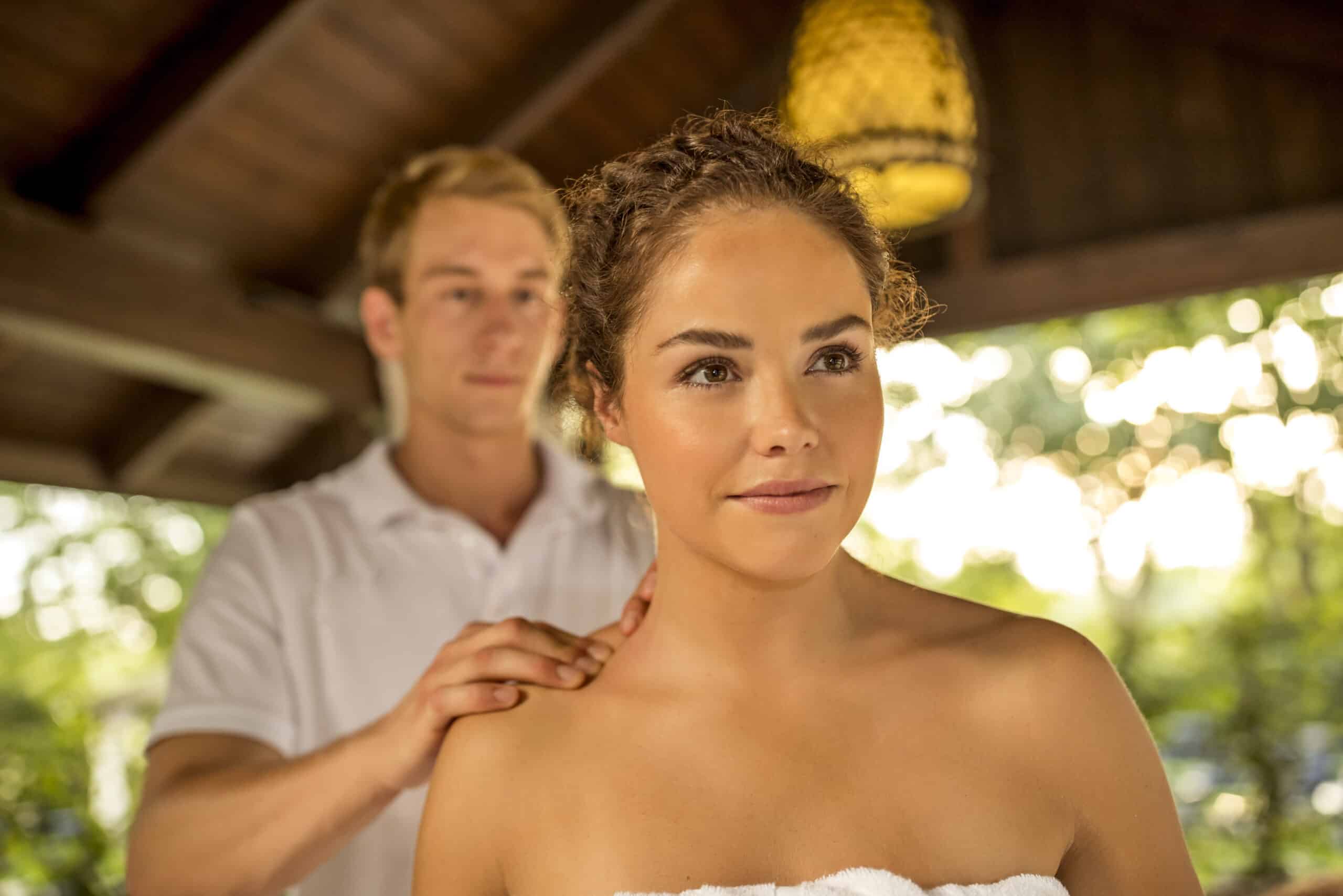 Eine Frau mit lockigem Haar, die in ein Handtuch gehüllt ist, erhält eine Schultermassage von einem Mann mit kurzen blonden Haaren und einem weißen Hemd. Sie befinden sich in einer Außenumgebung, im Hintergrund sind Holzbalken und Laub zu sehen. © Fotografie Tomas Rodriguez