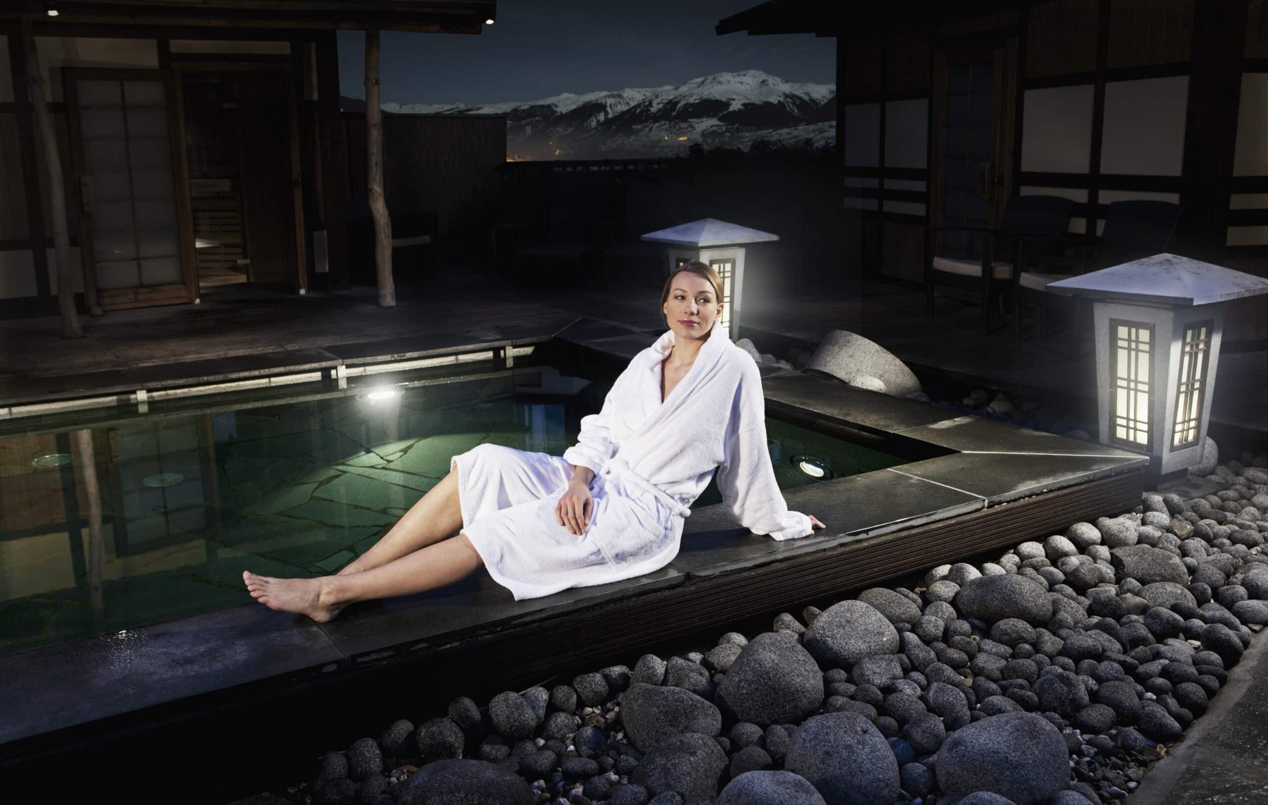 Eine Frau im weißen Bademantel sitzt nachts am Rand eines mit Steinen umrandeten Pools in einem Freiluft-Spa. Sie ist umgeben von glatten Steinen, Laternen und Holzkonstruktionen unter einem klaren Himmel mit schneebedeckten Bergen im Hintergrund. © Fotografie Tomas Rodriguez