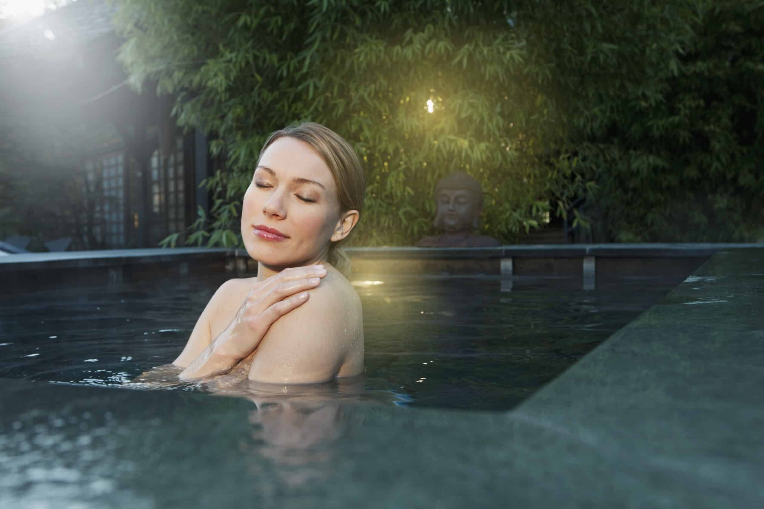 Eine Frau mit geschlossenen Augen entspannt sich in einer heißen Quelle oder einem Pool im Freien und hält sich dabei sanft die Schulter. Im Hintergrund sind üppiges Grün und eine Buddha-Statue zu sehen, die eine ruhige und heitere Atmosphäre schaffen. Sanftes Sonnenlicht fällt hinein und verstärkt die friedliche Stimmung. © Fotografie Tomas Rodriguez