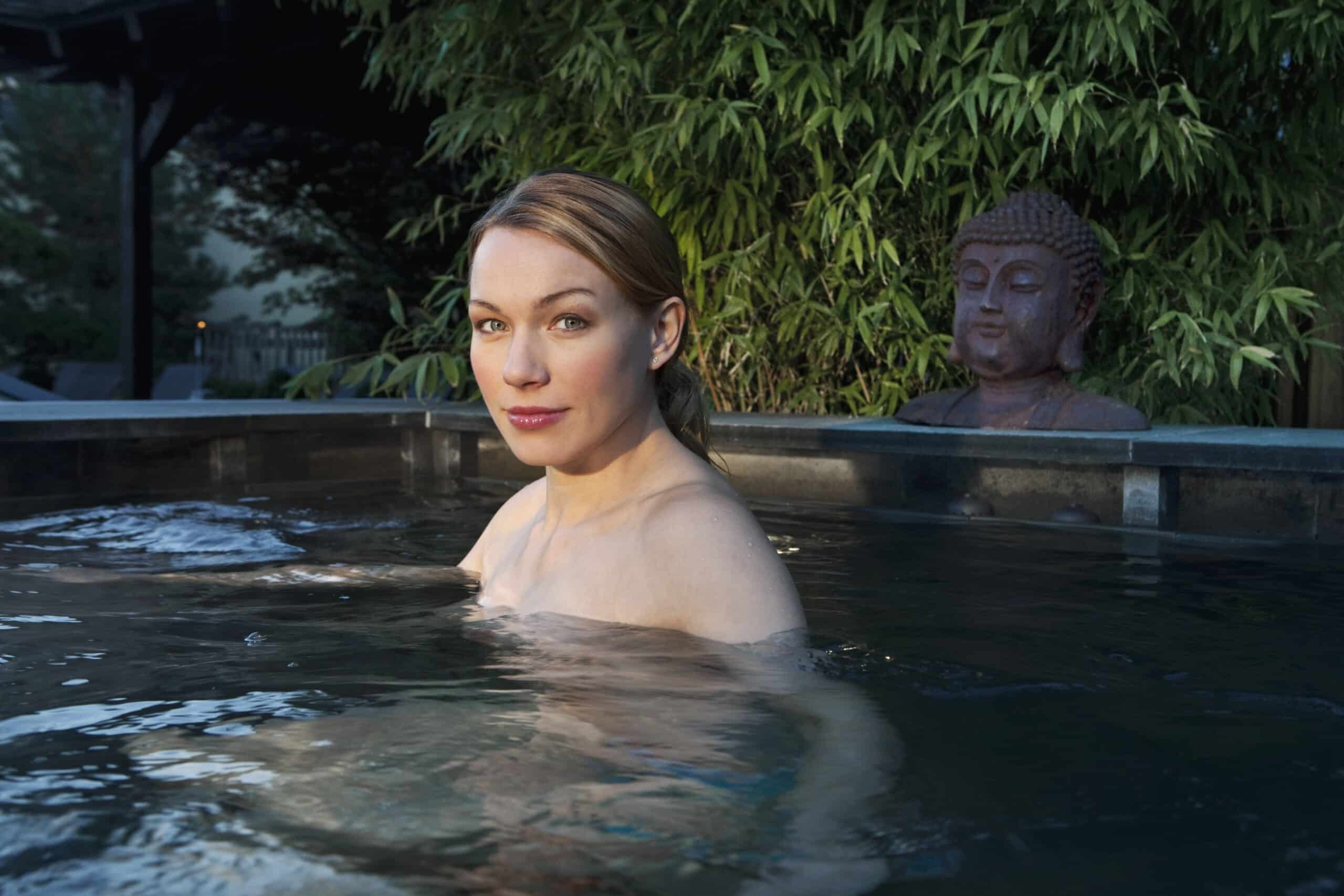 Eine Person mit hellbraunem Haar entspannt sich in einem Whirlpool im Freien. Das Wasser ist ruhig und im Hintergrund sind grüne Bambuspflanzen und eine Buddha-Statue zu sehen, die eine heitere Atmosphäre schaffen. © Fotografie Tomas Rodriguez
