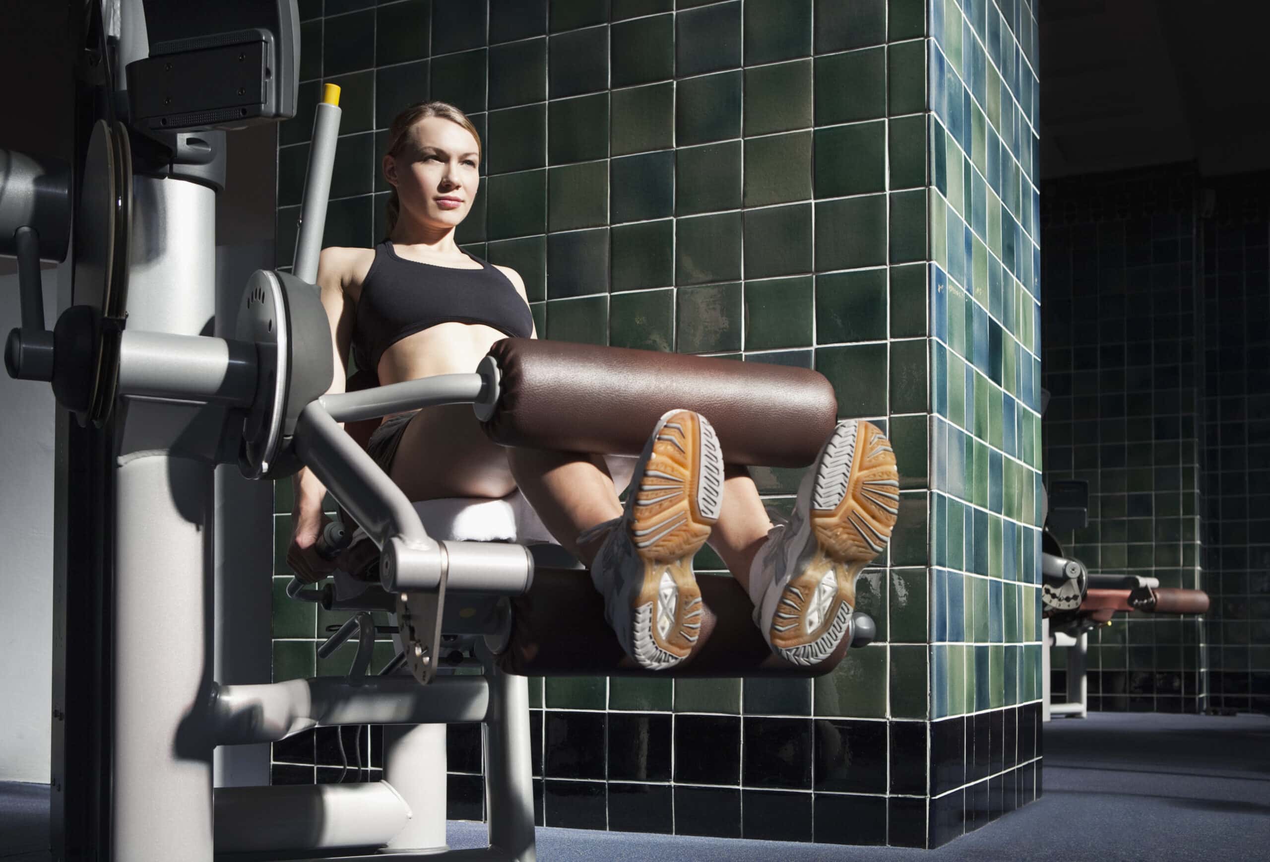 Eine Frau in Sportkleidung benutzt in einem Fitnessstudio eine Beinstreckermaschine. Sie stemmt Gewichte, wobei beide Beine nach vorne gestreckt sind. Im Hintergrund sind grün geflieste Wände zu sehen. © Fotografie Tomas Rodriguez