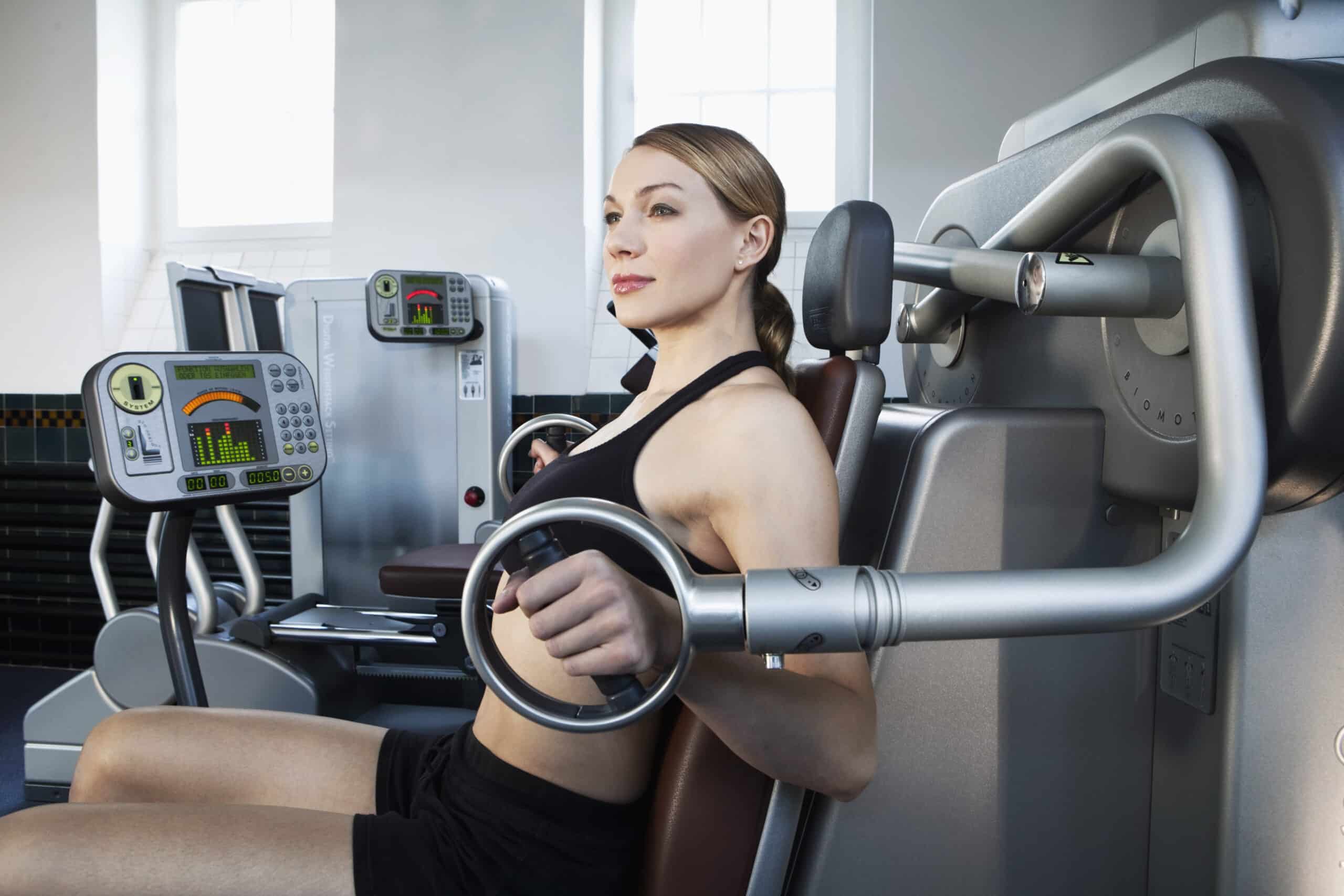 Eine Frau in Trainingskleidung trainiert konzentriert an einer Bankpresse im Fitnessstudio. Im Hintergrund ist ein weiteres Trainingsgerät mit digitaler Anzeige und Bedienelementen zu sehen. Das Fitnessstudio ist durch ein Fenster mit natürlichem Licht gut beleuchtet. © Fotografie Tomas Rodriguez