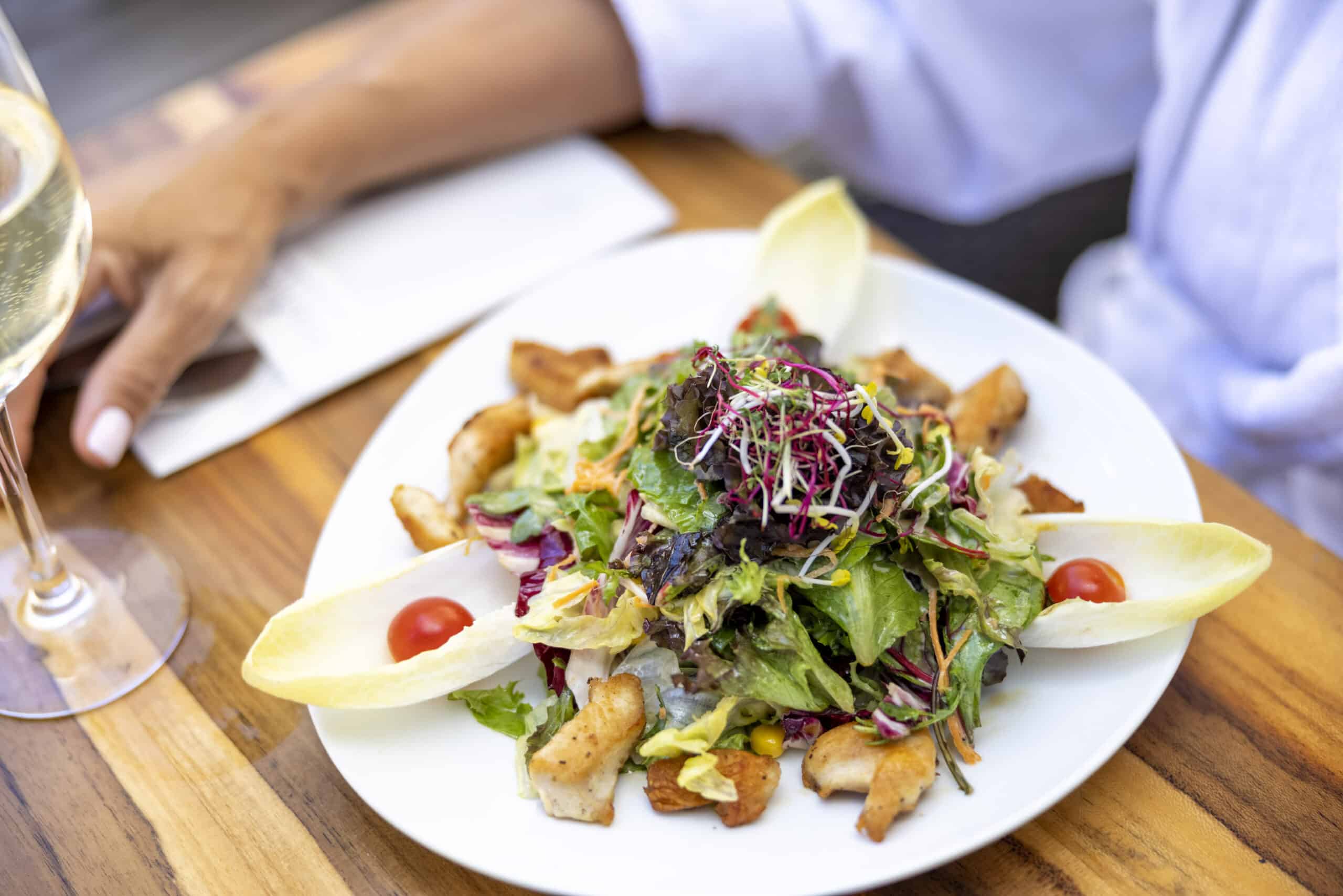 Eine Person sitzt an einem Holztisch und hat einen weißen Teller mit frischem Salat, gemischten Blattsalaten, Kirschtomaten, Croutons und Endivienblättern. Links neben dem Teller steht ein Glas Weißwein. In einer Hand hält die Person eine Speisekarte oder Broschüre. © Fotografie Tomas Rodriguez
