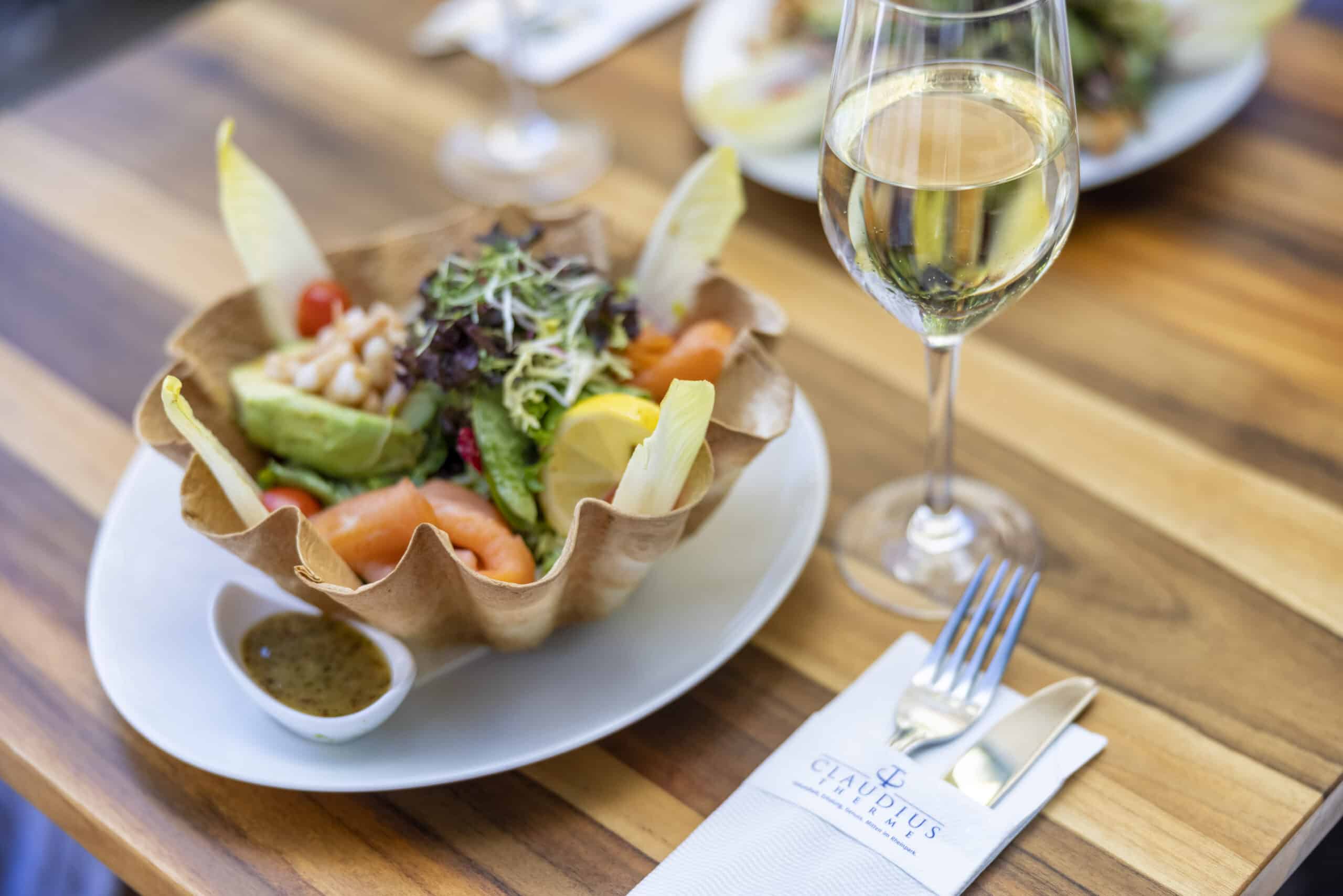 Ein gedeckter Holztisch mit einem Glas Weißwein und Gabel und Messer auf einer weißen Serviette, auf der „Cayman Islands“ aufgedruckt ist. Als Hauptspeise gibt es einen bunten Salat in einer knusprigen Taco-Schale, dazu Dressing in einer kleinen Schüssel. © Fotografie Tomas Rodriguez