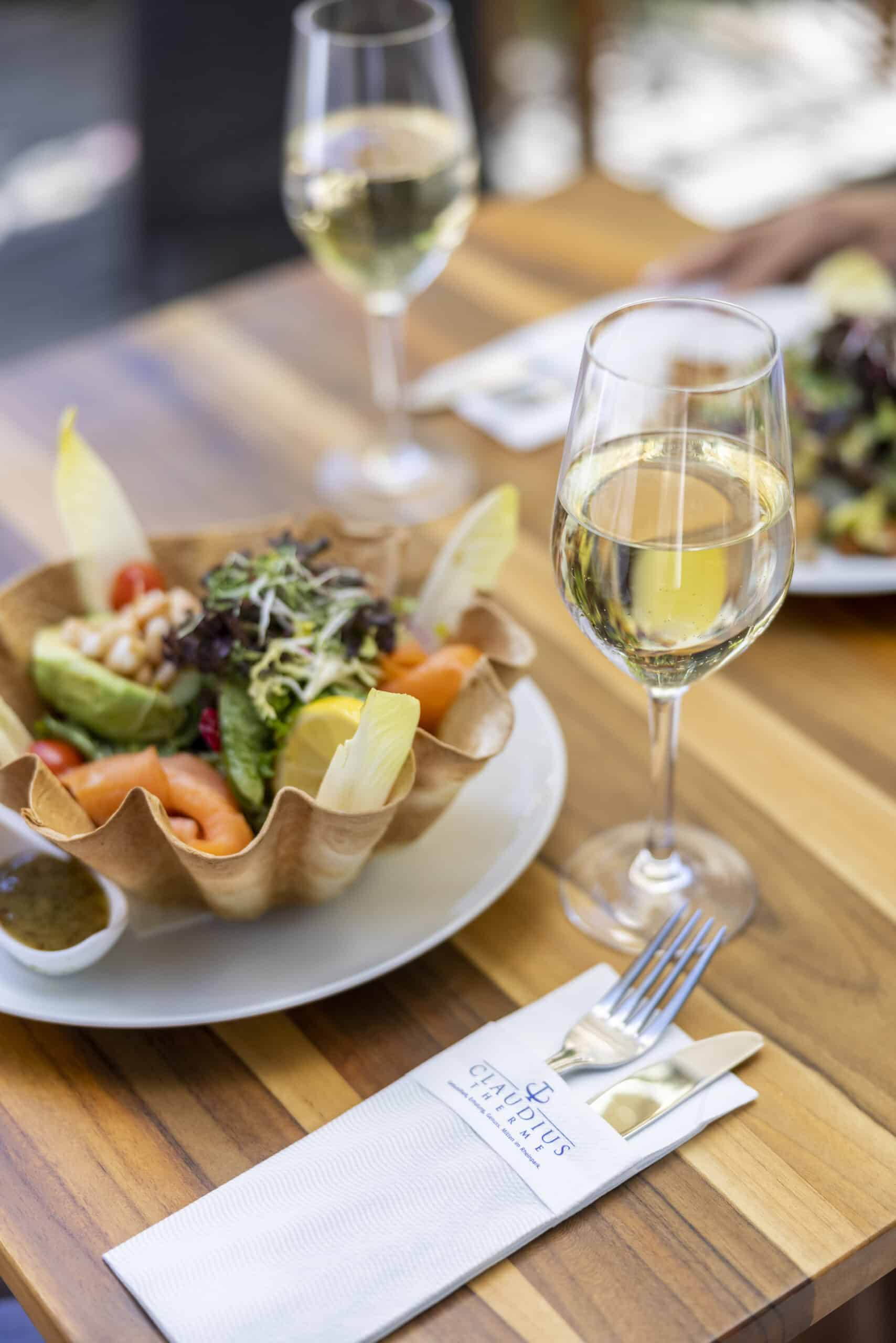Ein für zwei Personen gedeckter Holztisch bietet zwei Gläser Weißwein, einen bunten Salat in einer Schüssel aus knuspriger Schale und eine weiße Serviette mit Logo, Gabel und Messer. Der Salat besteht aus Garnelen, Avocado, Kirschtomaten, Blattgemüse und Endivienblättern. © Fotografie Tomas Rodriguez