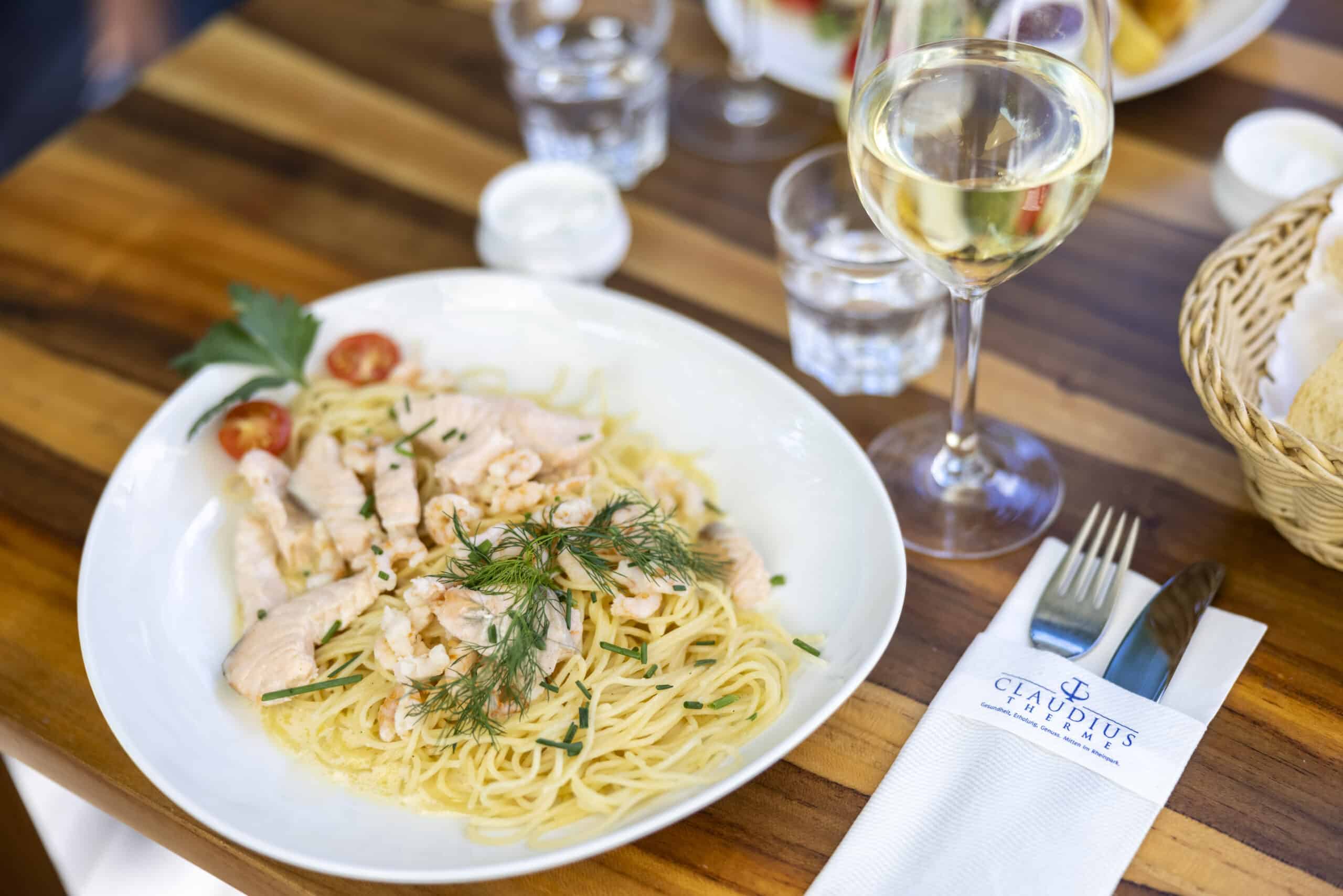 Ein Teller Spaghetti mit Garnelen, garniert mit frischen Kräutern. Neben dem Teller steht ein mit Weißwein gefülltes Weinglas, ein Gabel- und Messerset auf einer Markenserviette und ein Weidenkorb mit Brot. Der Tisch ist mit weiteren Gläsern und einer kleinen Schale gedeckt. © Fotografie Tomas Rodriguez