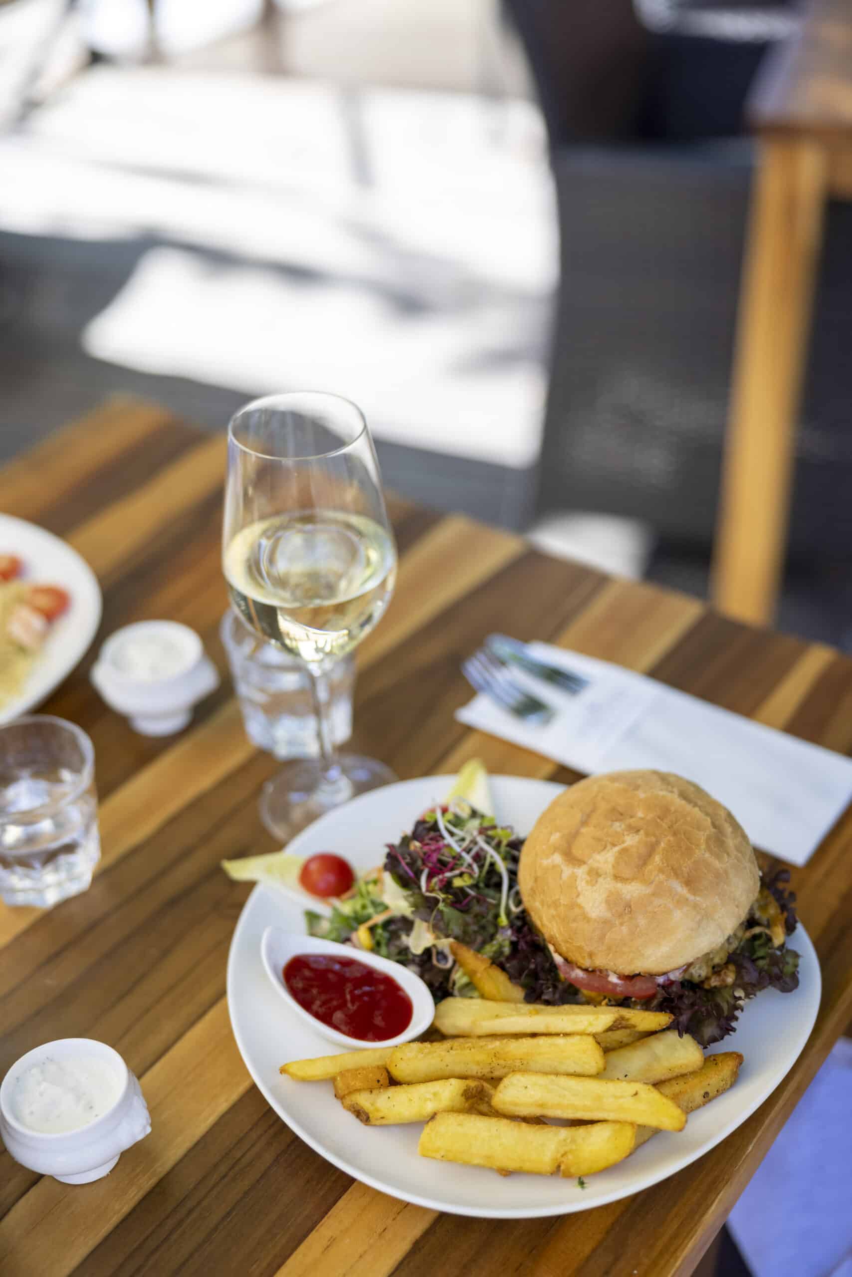 Ein Teller mit Essen auf einem Holztisch umfasst einen Burger mit frischem Gemüse, dick geschnittene Pommes, Ketchup und einen kleinen Salat. Ein Glas Weißwein, eine Gabel und ein Messer stehen neben dem Teller, zusammen mit kleinen Gewürzbehältern. Der Hintergrund ist leicht unscharf. © Fotografie Tomas Rodriguez