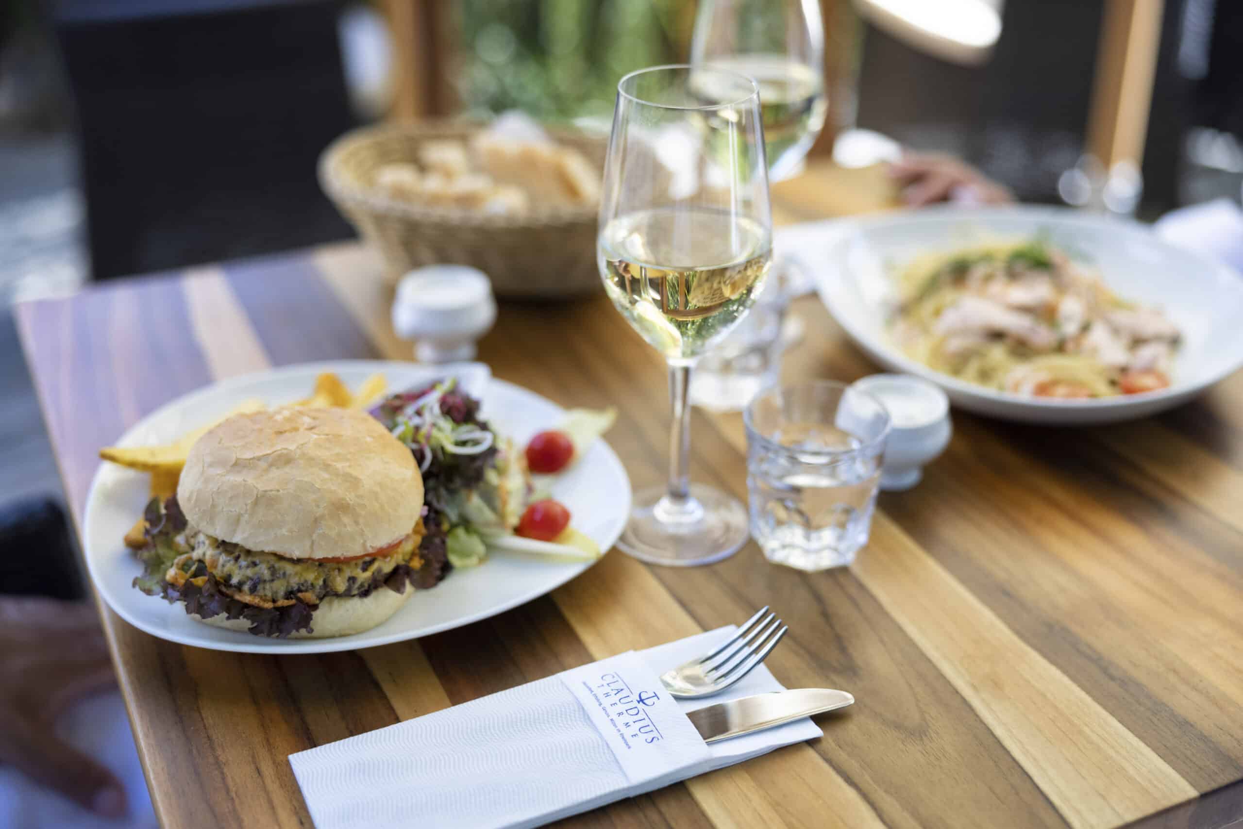 Ein für eine Mahlzeit gedeckter Holztisch: Auf einem Teller liegt ein Cheeseburger mit Pommes und einem gemischten Salat, im Hintergrund ein Nudelgericht, im Hintergrund ein Korb mit Brot, ein Glas Weißwein, ein Wasserglas und darauf eine gefaltete Serviette mit Gabel und Messer. © Fotografie Tomas Rodriguez