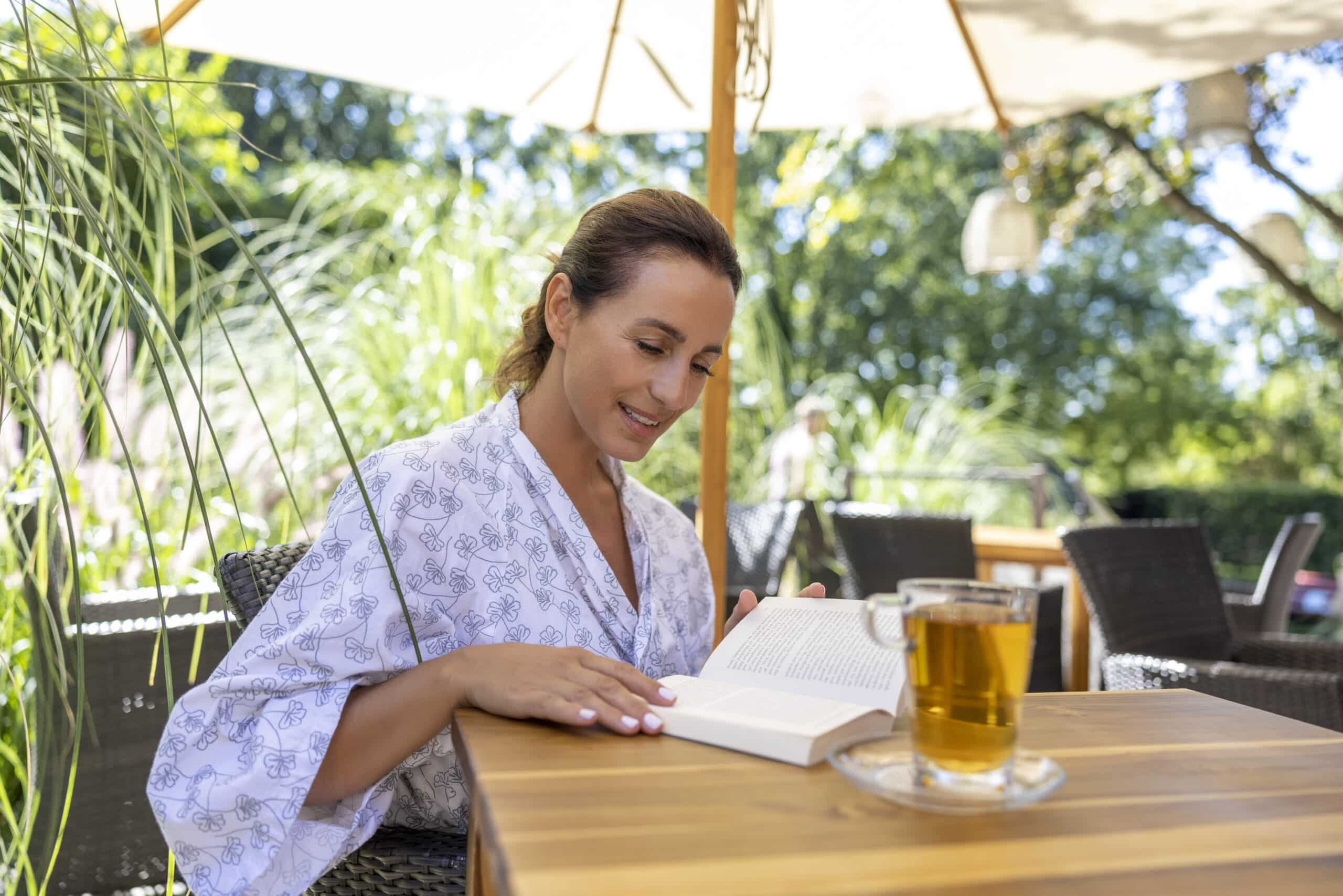 Eine Frau sitzt an einem Holztisch im Freien und liest ein Buch. Sie trägt einen hellen, gemusterten Bademantel oder Freizeitkleidung. Vor ihr steht auf dem Tisch eine Tasse Tee. Sie steht unter einem großen Sonnenschirm mit Grünpflanzen im Hintergrund. © Fotografie Tomas Rodriguez