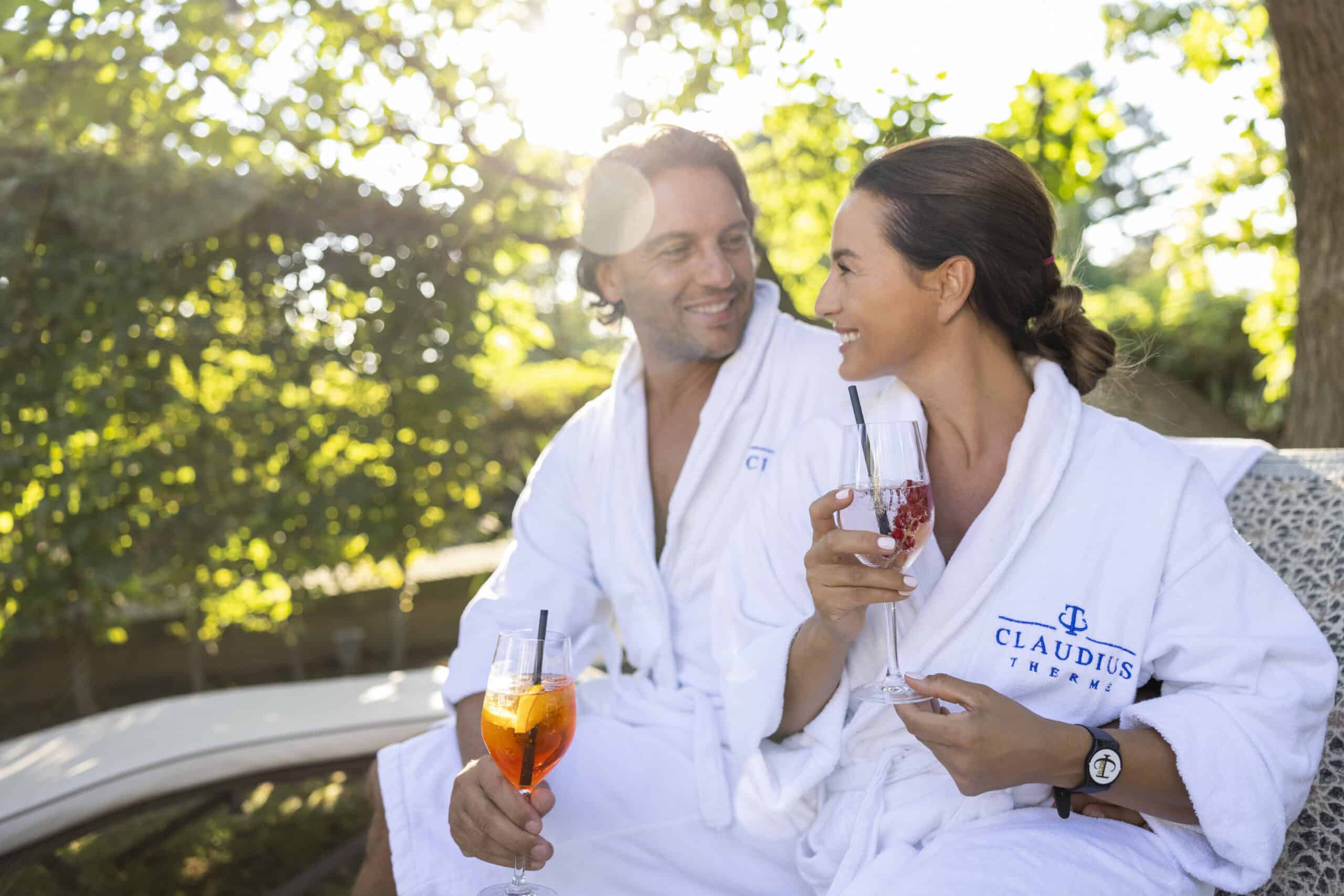 Ein Mann und eine Frau in weißen Roben sitzen draußen und lächeln sich mit Cocktailgläsern in den Händen an. Auf die Roben ist „Claudius Therme“ gestickt. Sie sind von üppigem Grün umgeben und Sonnenlicht fällt durch die Bäume. © Fotografie Tomas Rodriguez