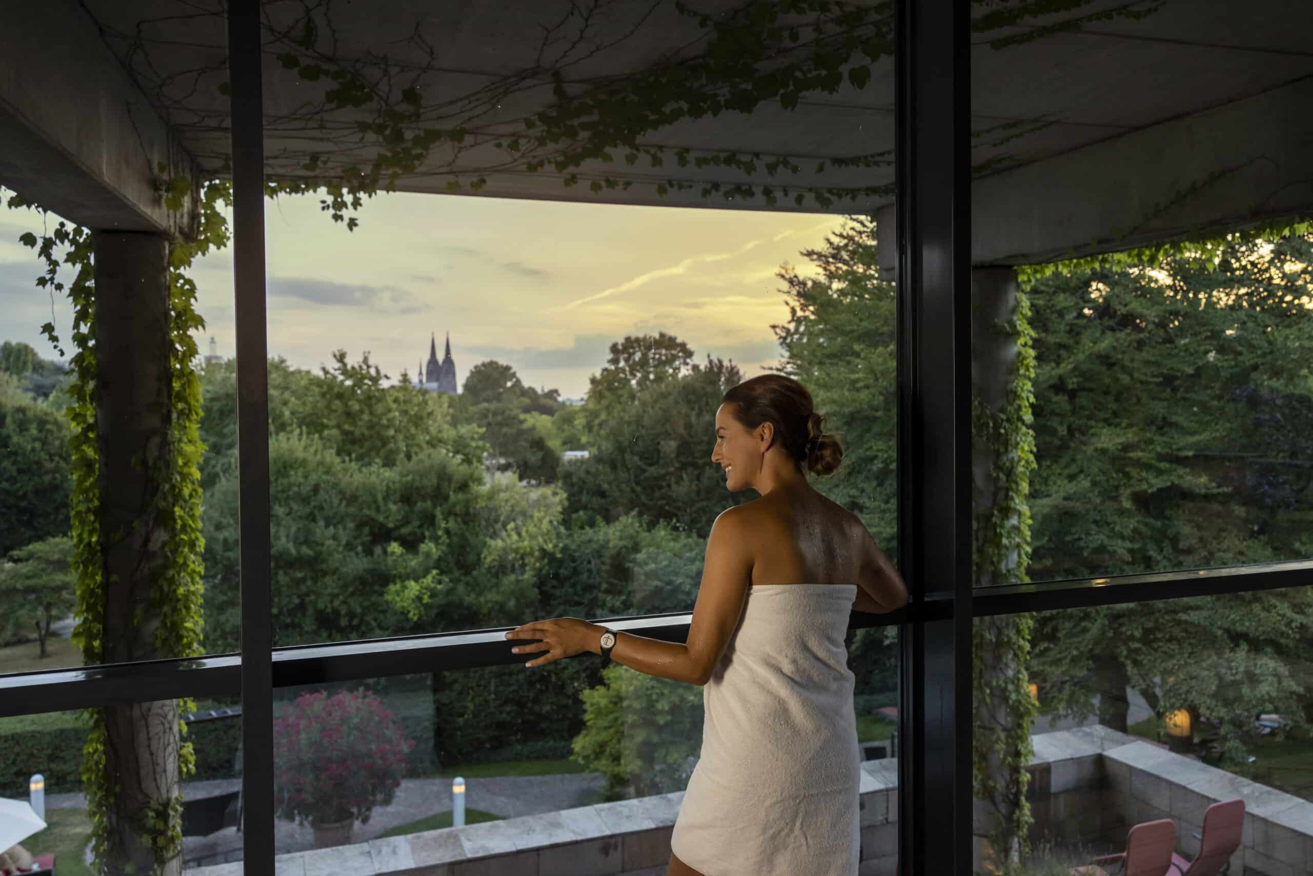 Eine Frau steht in ein Handtuch gehüllt auf einem Balkon mit efeubewachsenen Säulen. Ihr Blick fällt auf eine ruhige, baumreiche Landschaft, in der Ferne ist ein Kirchturm zu sehen, der Himmel ist in der Abenddämmerung teilweise bewölkt. © Fotografie Tomas Rodriguez