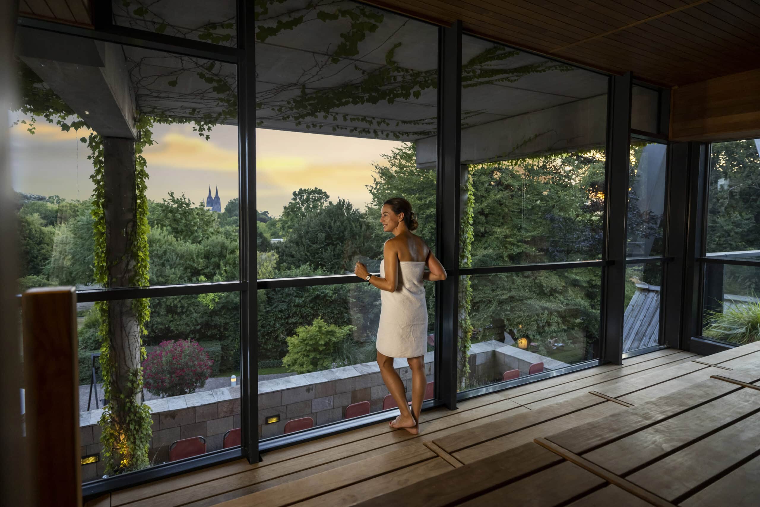 Eine Frau in einem Handtuch steht auf einem Innenbalkon und blickt durch große Fenster auf eine üppige, grüne Landschaft im Sonnenuntergang. Der Raum hat Holzböden und eine begrünte Außenwand, im Hintergrund ist ein Kirchturm zu sehen. © Fotografie Tomas Rodriguez