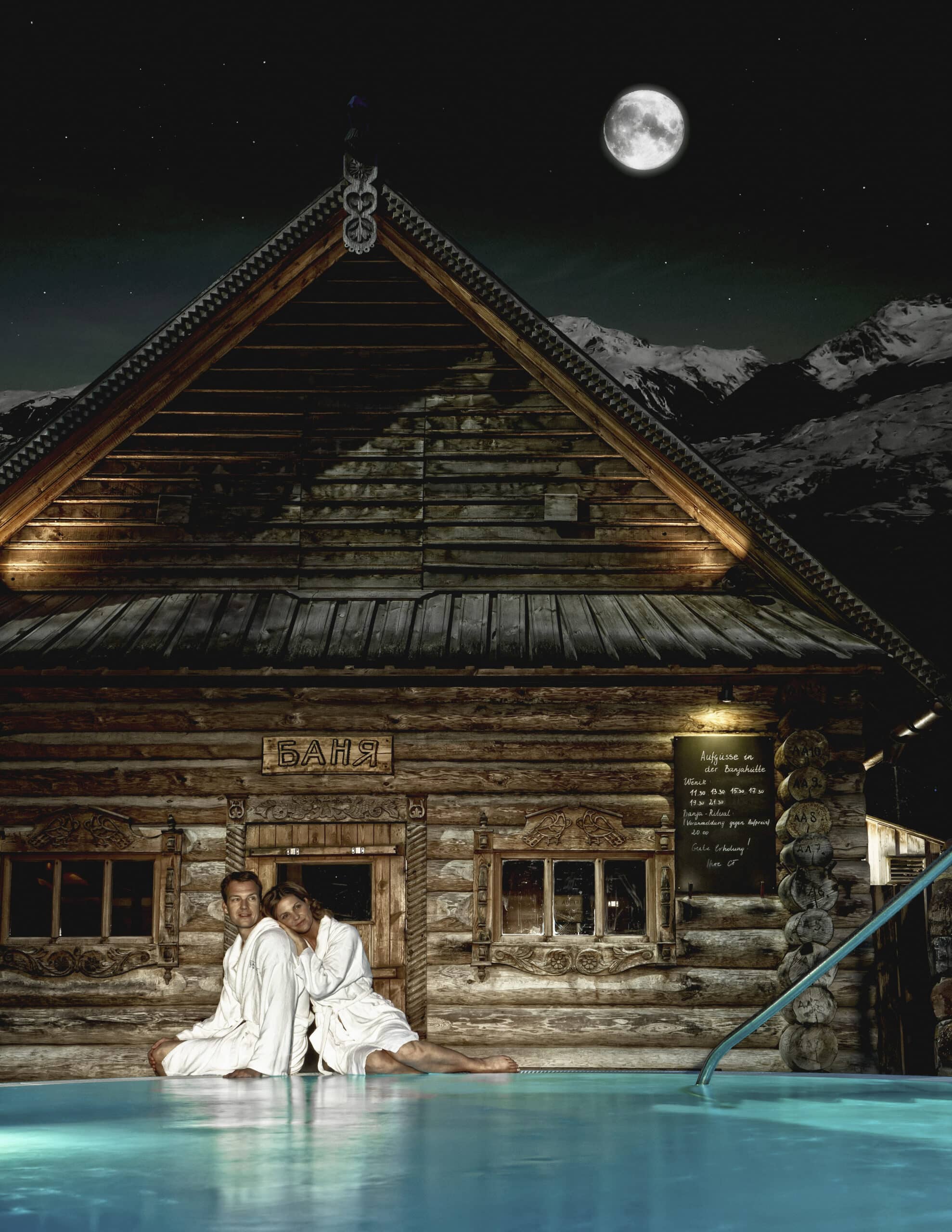 Ein Mann und eine Frau, beide in weiße Gewänder gekleidet, sitzen bei Vollmond auf den Holzstufen einer rustikalen Blockhütte. Die Hütte hat ein Schild mit kyrillischer Schrift und ist von schneebedeckten Bergen umgeben. Ein Teich im Vordergrund spiegelt die Szene wider. © Fotografie Tomas Rodriguez