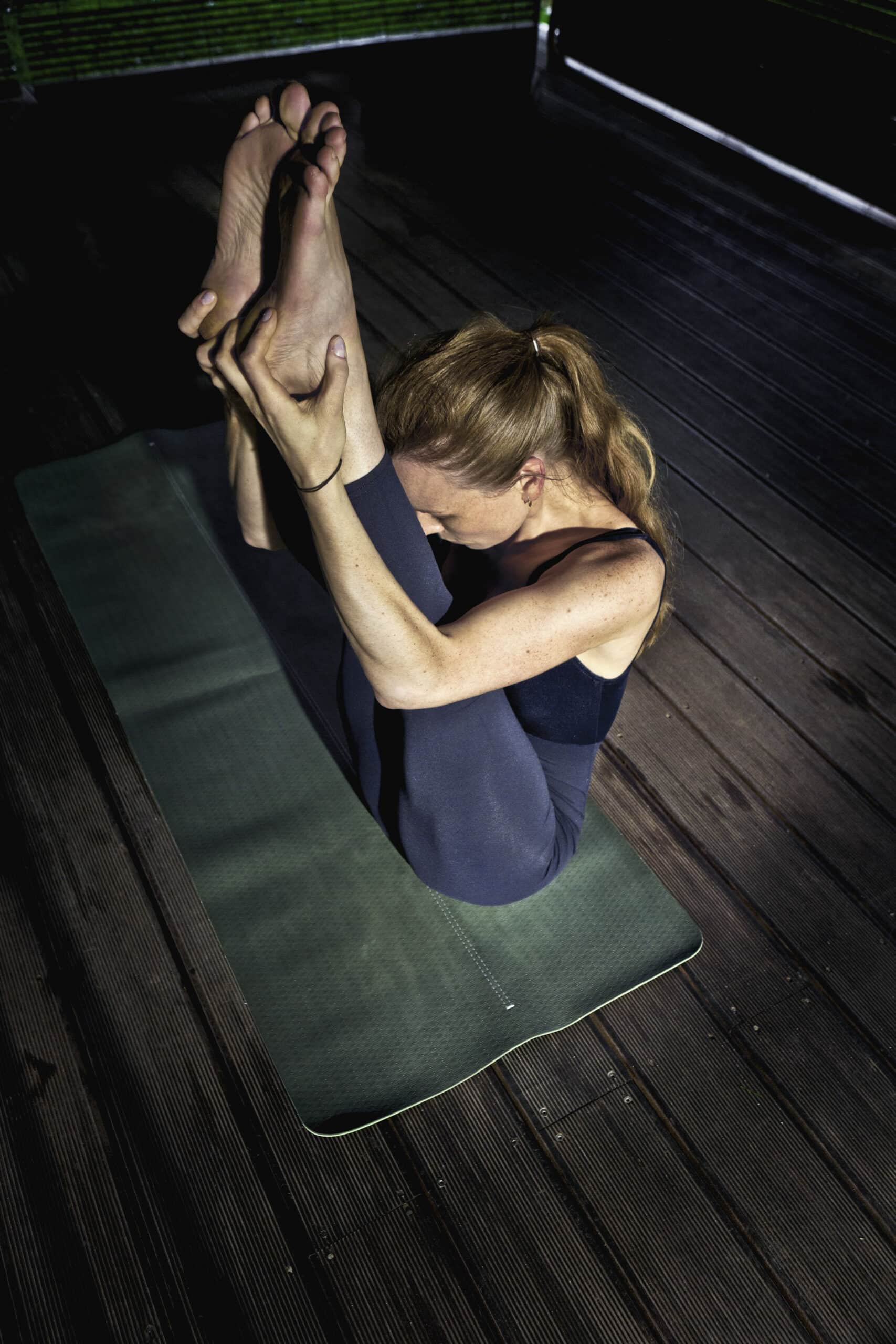 Eine Frau mit blondem Haar führt eine Yoga-Pose auf einer grünen Matte aus. Sie sitzt, die Beine sind nach oben gestreckt und ihre Füße sind mit den Händen umschlossen. Die Umgebung scheint ein schwach beleuchteter Holzboden zu sein. Sie trägt dunkle Sportkleidung. © Fotografie Tomas Rodriguez