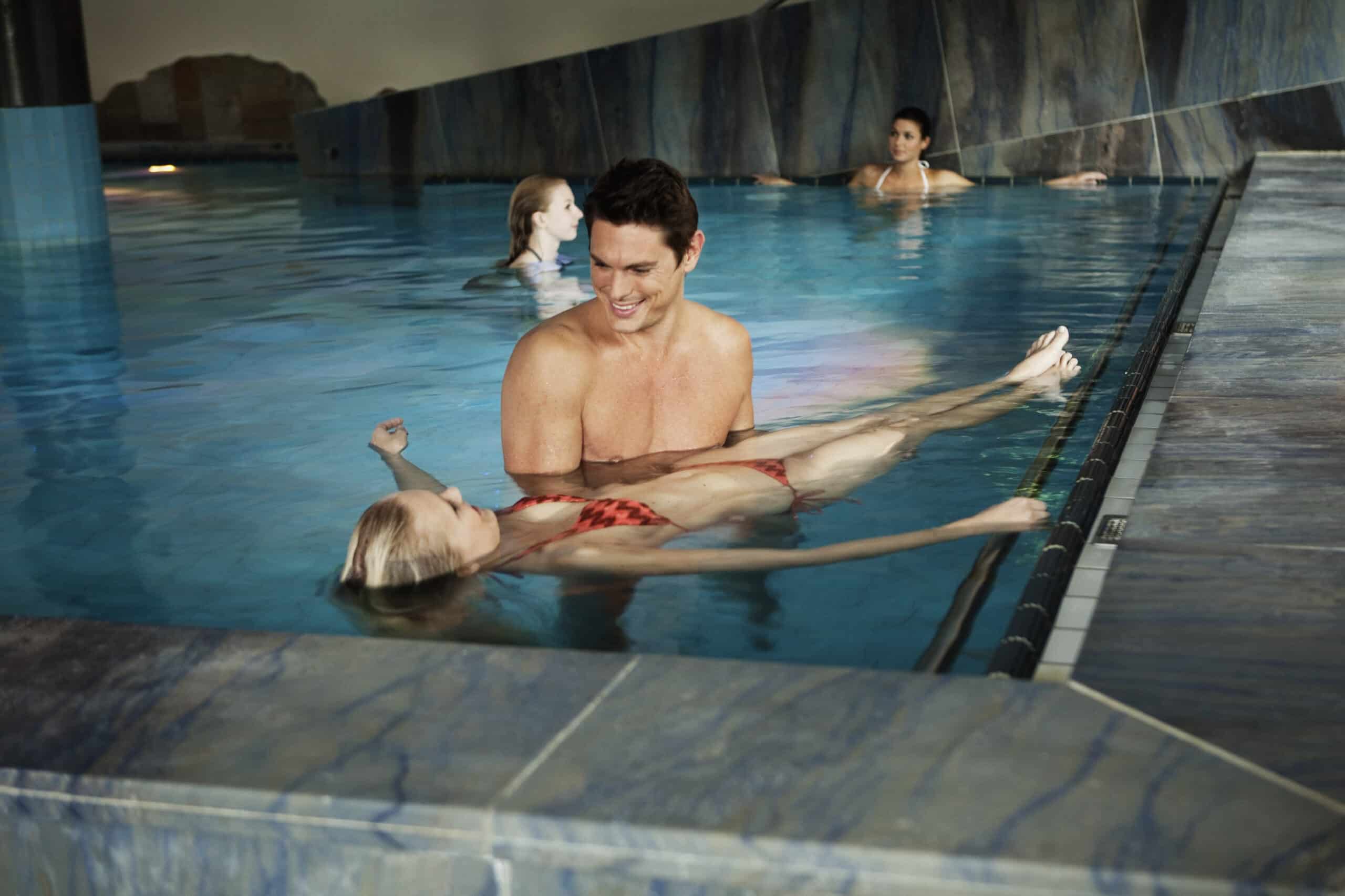 Ein Mann hält eine Frau, die auf dem Rücken in einem Hallenbad schwimmt. Beide tragen Badebekleidung und lächeln sich an. Im Hintergrund schwimmen zwei weitere Personen im Pool. Der Poolbereich ist modern gestaltet und mit blauen Fliesen versehen. © Fotografie Tomas Rodriguez