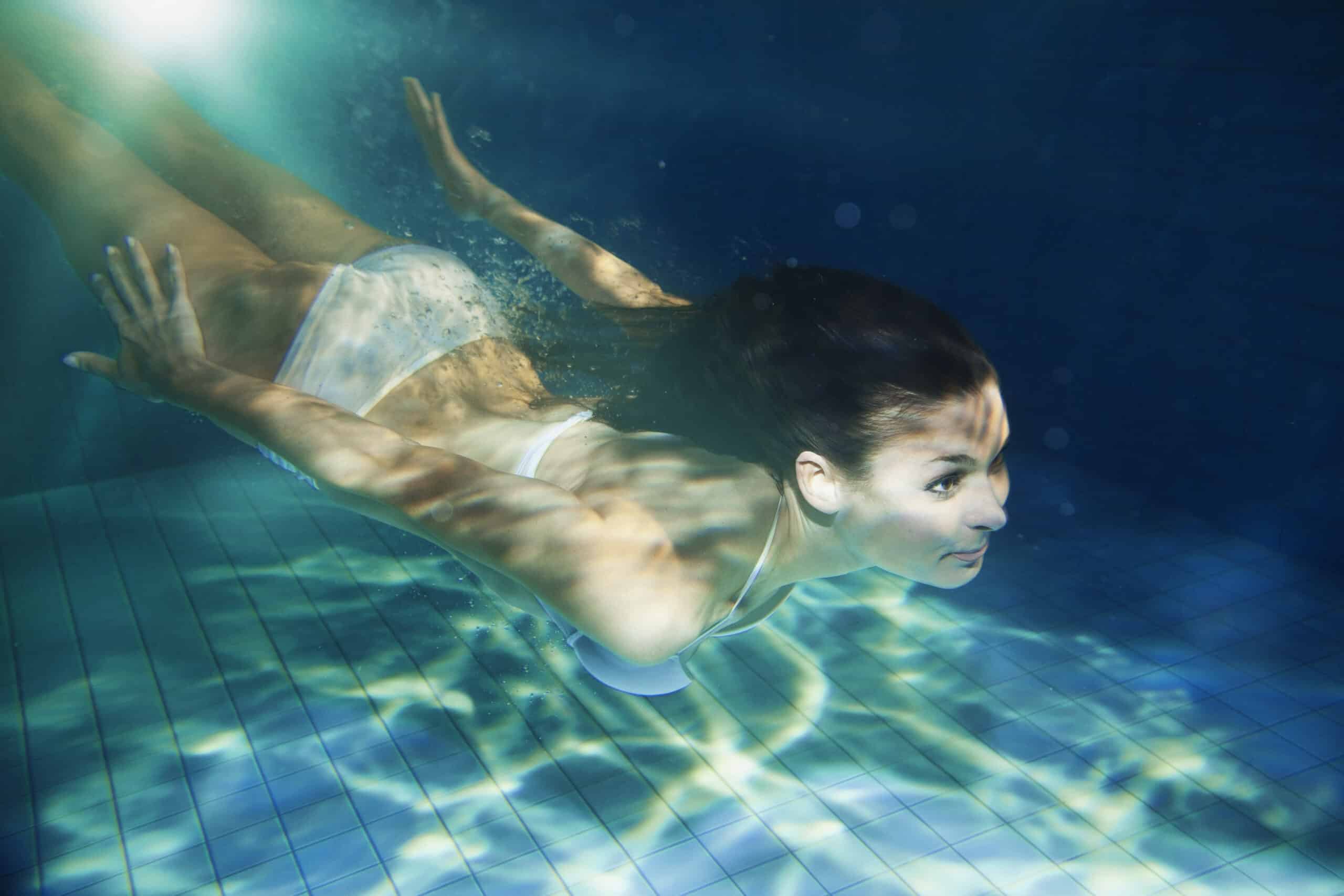 Eine Frau in einem weißen Badeanzug schwimmt unter Wasser in einem Pool. Ihre Arme sind nach hinten ausgestreckt und ihr Körper gleitet stromlinienförmig durch das Wasser. Sonnenlicht fällt durch das Wasser und wirft Muster auf ihren Körper und den Poolboden. © Fotografie Tomas Rodriguez