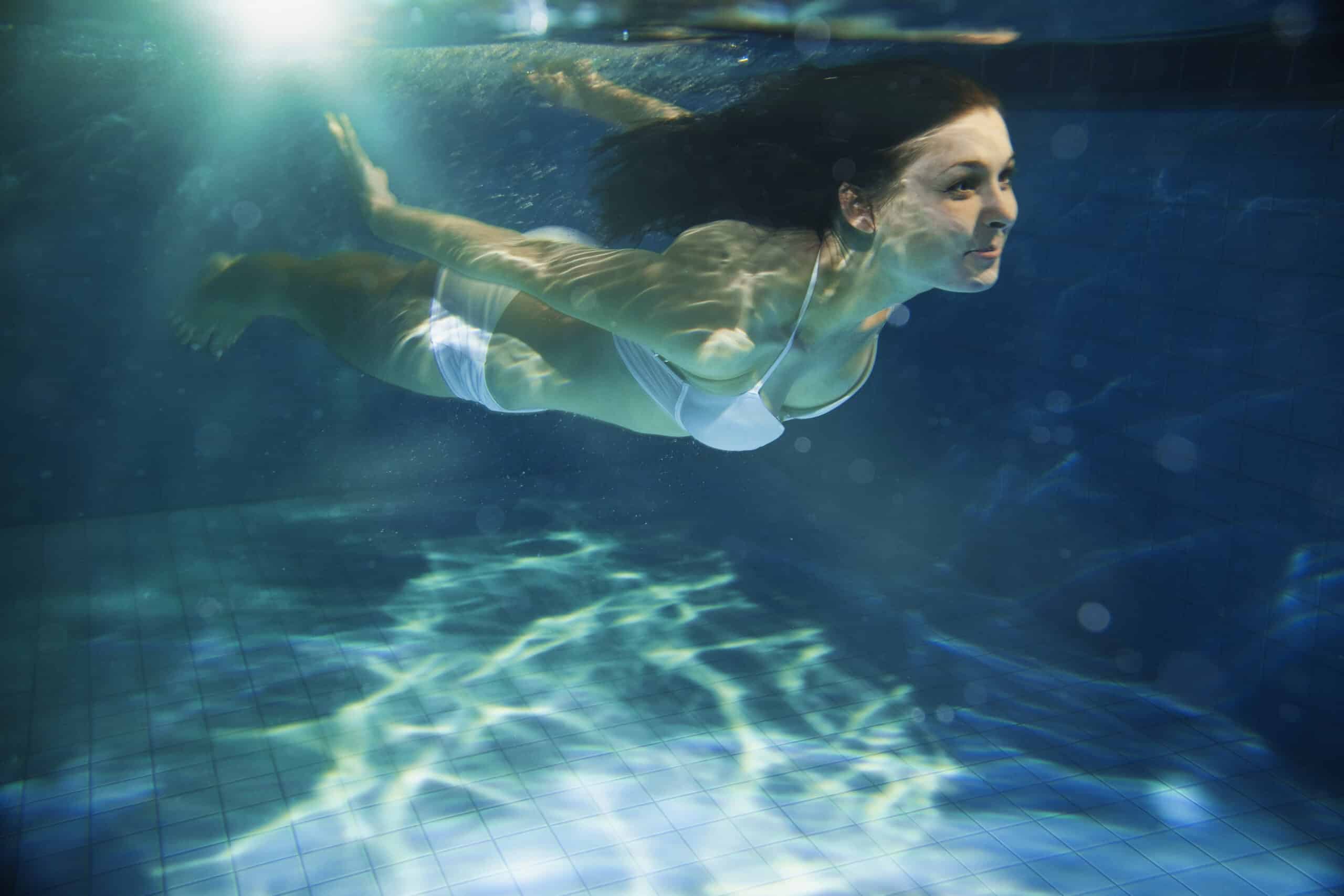 Eine Frau mit langen Haaren schwimmt unter Wasser in einem blau gefliesten Pool. Sie trägt einen weißen Bikini und das Sonnenlicht fällt durch das Wasser und erzeugt gesprenkelte Muster auf dem Poolboden und ihrer Haut. © Fotografie Tomas Rodriguez