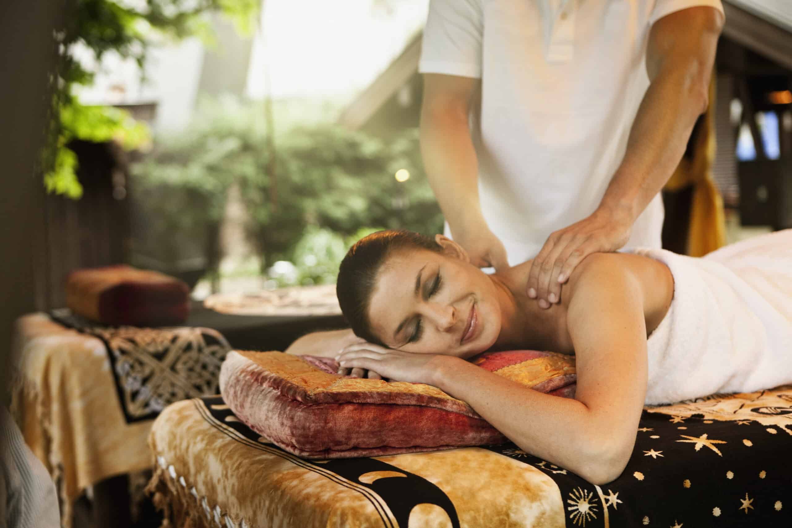 Eine Frau liegt mit dem Gesicht nach unten auf einem Massagetisch und ist in ein Handtuch gewickelt. Eine Person gibt ihr eine Schultermassage. Die Umgebung ist ruhig, mit natürlichem Licht und im Hintergrund sind Grünpflanzen zu sehen. Die Frau wirkt entspannt und zufrieden. © Fotografie Tomas Rodriguez
