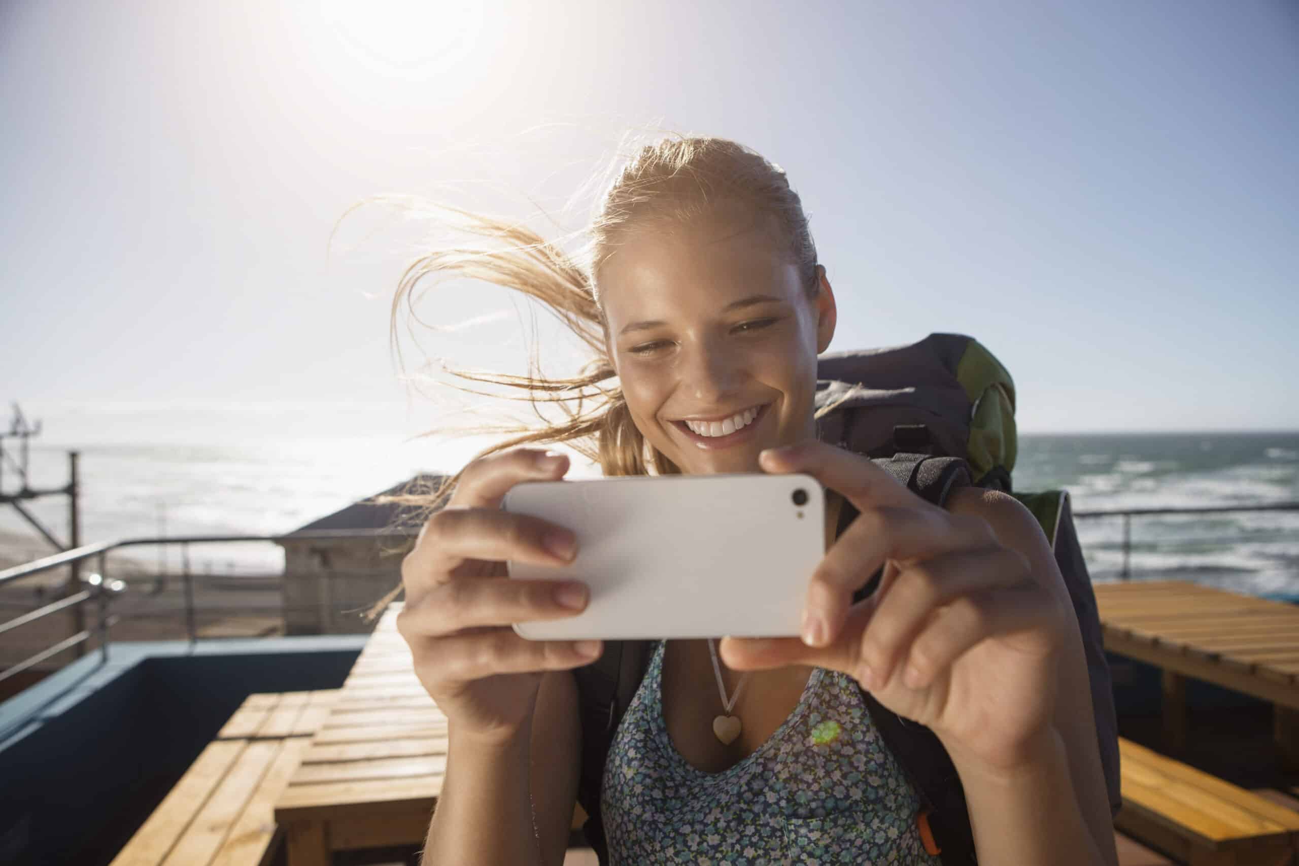 Eine lächelnde Person mit langen, im Wind wehenden Haaren macht ein Foto mit einem Smartphone. Sie stehen an einem sonnigen Tag draußen am Meer, im Hintergrund sind Holztische und Geländer zu sehen. Der Himmel ist klar und die Sonne scheint hell. © Fotografie Tomas Rodriguez