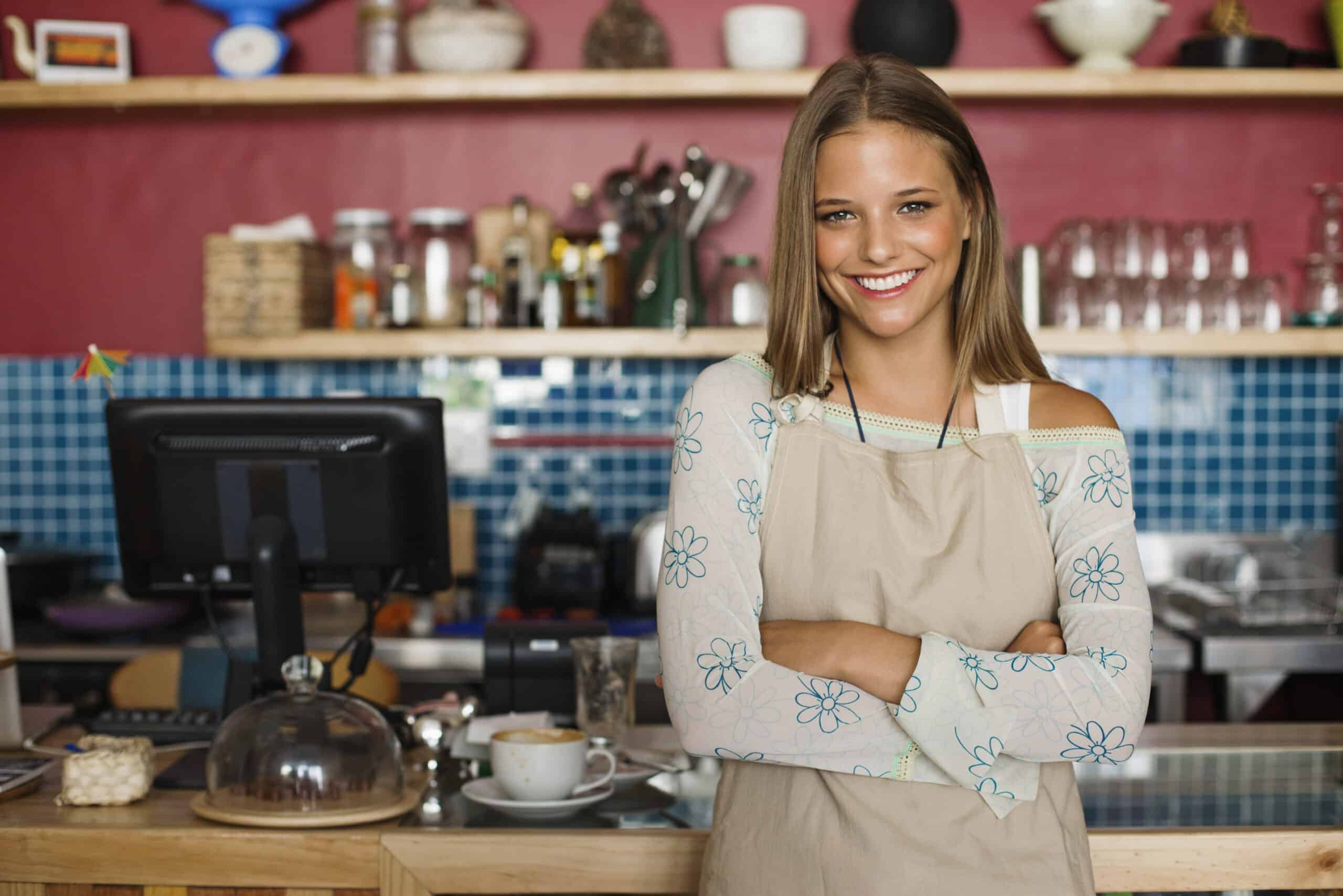 Eine lächelnde Frau mit langen Haaren steht hinter der Theke eines Cafés. Sie trägt eine beige Schürze über einem geblümten Hemd und hat die Arme verschränkt. Im Hintergrund sind Regale mit verschiedenen Artikeln, darunter Flaschen und Gläser, und ein Kassenterminal zu sehen. © Fotografie Tomas Rodriguez