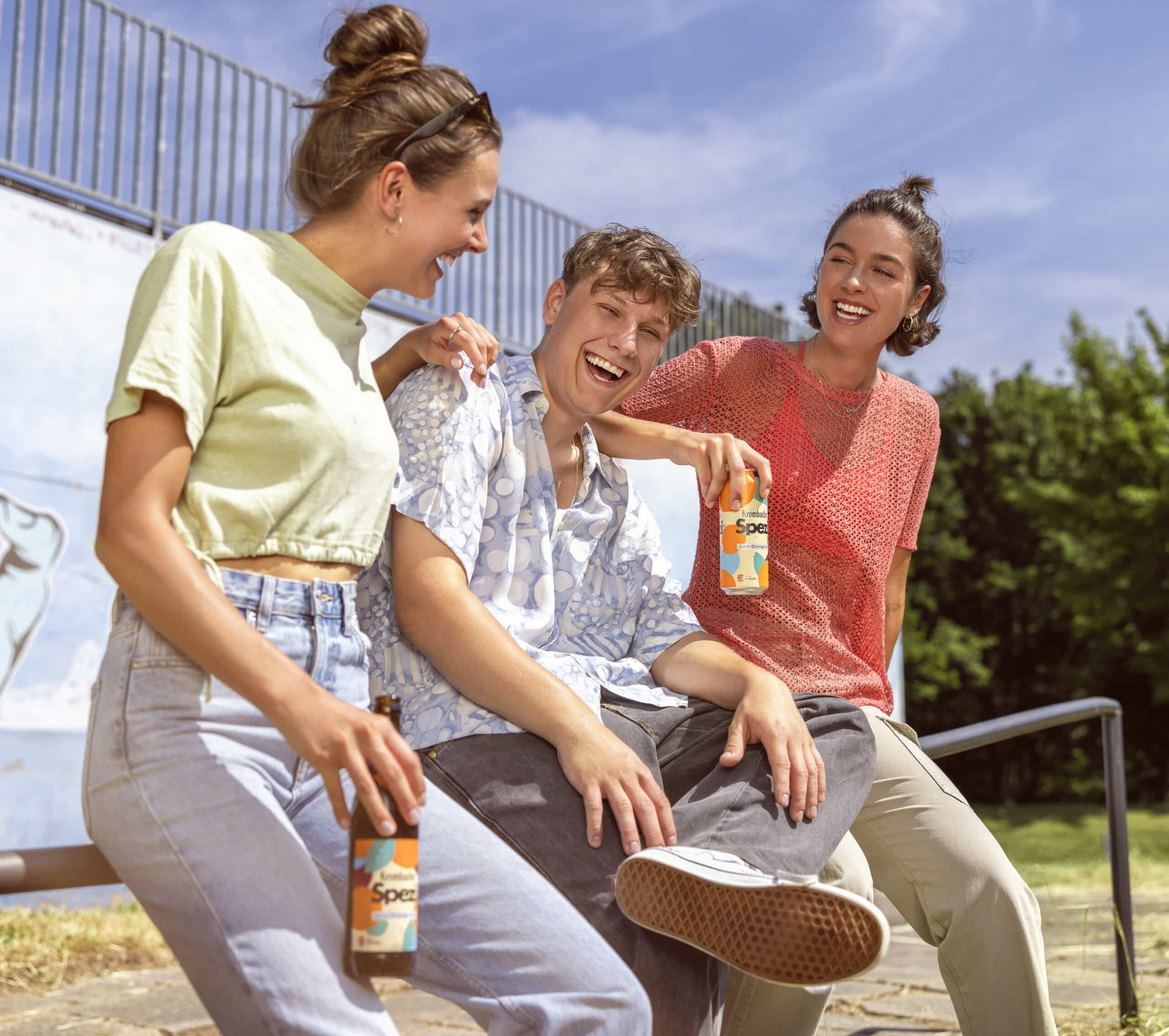 Drei Freunde sitzen an einem sonnigen Tag draußen auf einem Geländer, lachen und halten Flaschen mit Spezi-Limonade in den Händen. Die Gruppe wirkt entspannt und gut gelaunt und genießt gemeinsam ihre Getränke. Im Hintergrund sind Bäume und eine Skaterampe zu sehen. © Fotografie Tomas Rodriguez
