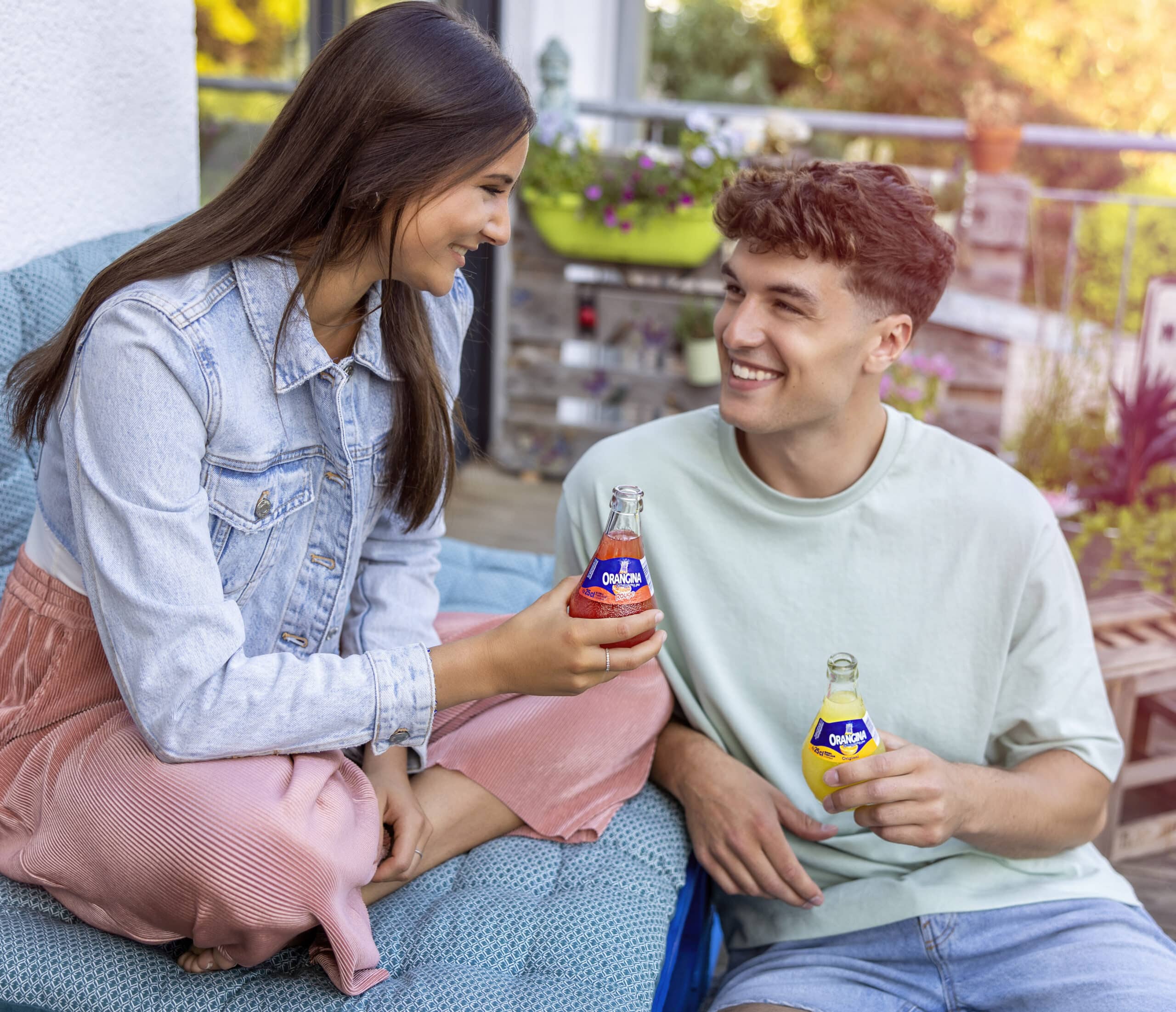 Eine Frau und ein Mann sitzen im Freien, lächeln sich an und halten Flaschen Olé-Limonade in den Händen. Die Frau trägt eine Jeansjacke und eine rosa Hose, der Mann ein hellgrünes Hemd und Shorts. Sie wirken entspannt und glücklich in dieser ungezwungenen Atmosphäre. © Fotografie Tomas Rodriguez