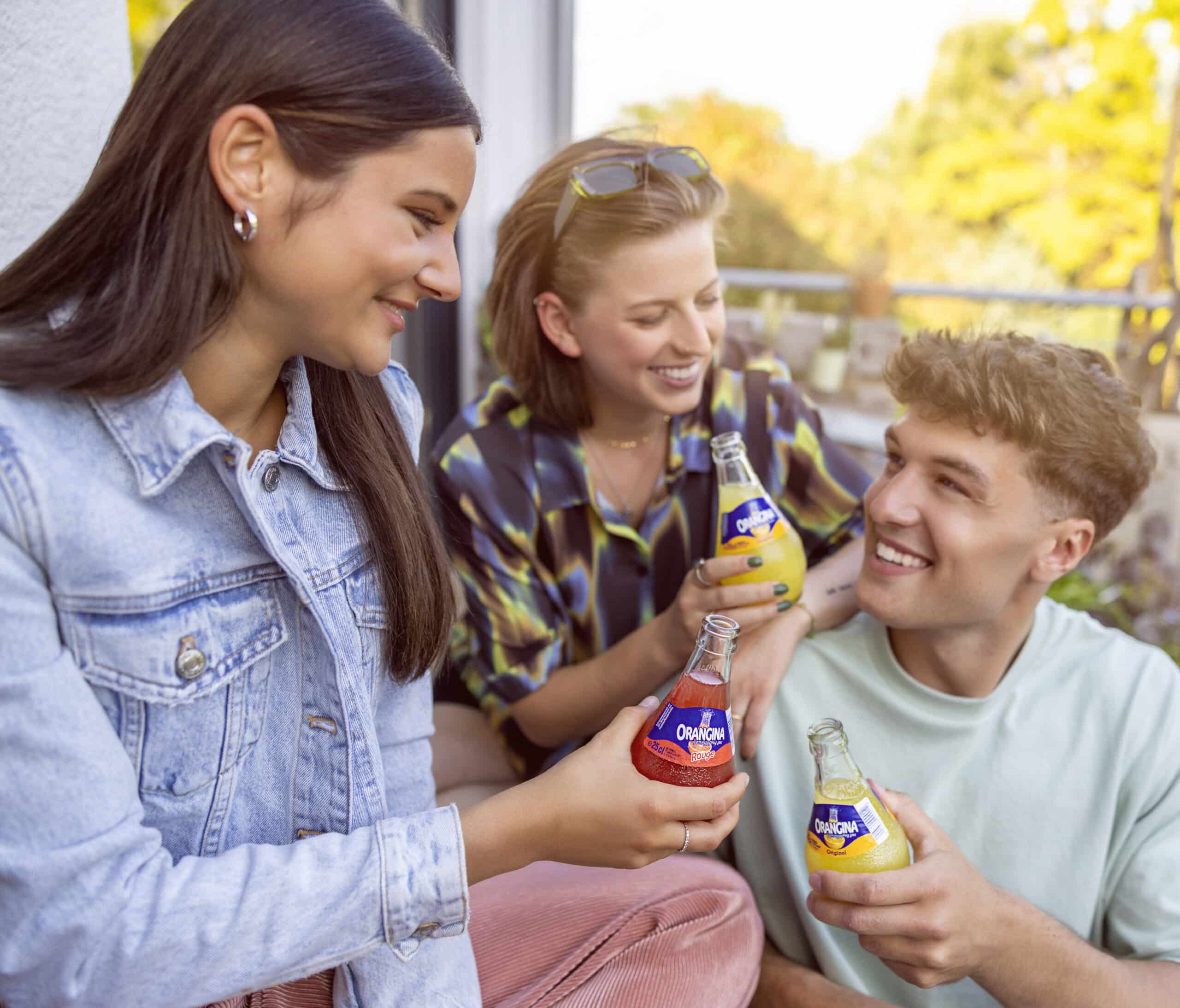Drei junge Erwachsene, eine Frau mit langen dunklen Haaren in einer Jeansjacke, eine andere mit kurzen blonden Haaren und einer Sonnenbrille auf dem Kopf in einem karierten Hemd und ein Mann mit lockigem Haar in einem hellgrünen Hemd, lächeln und halten Getränkeflaschen in der Hand, während sie sich im Freien unterhalten. © Fotografie Tomas Rodriguez