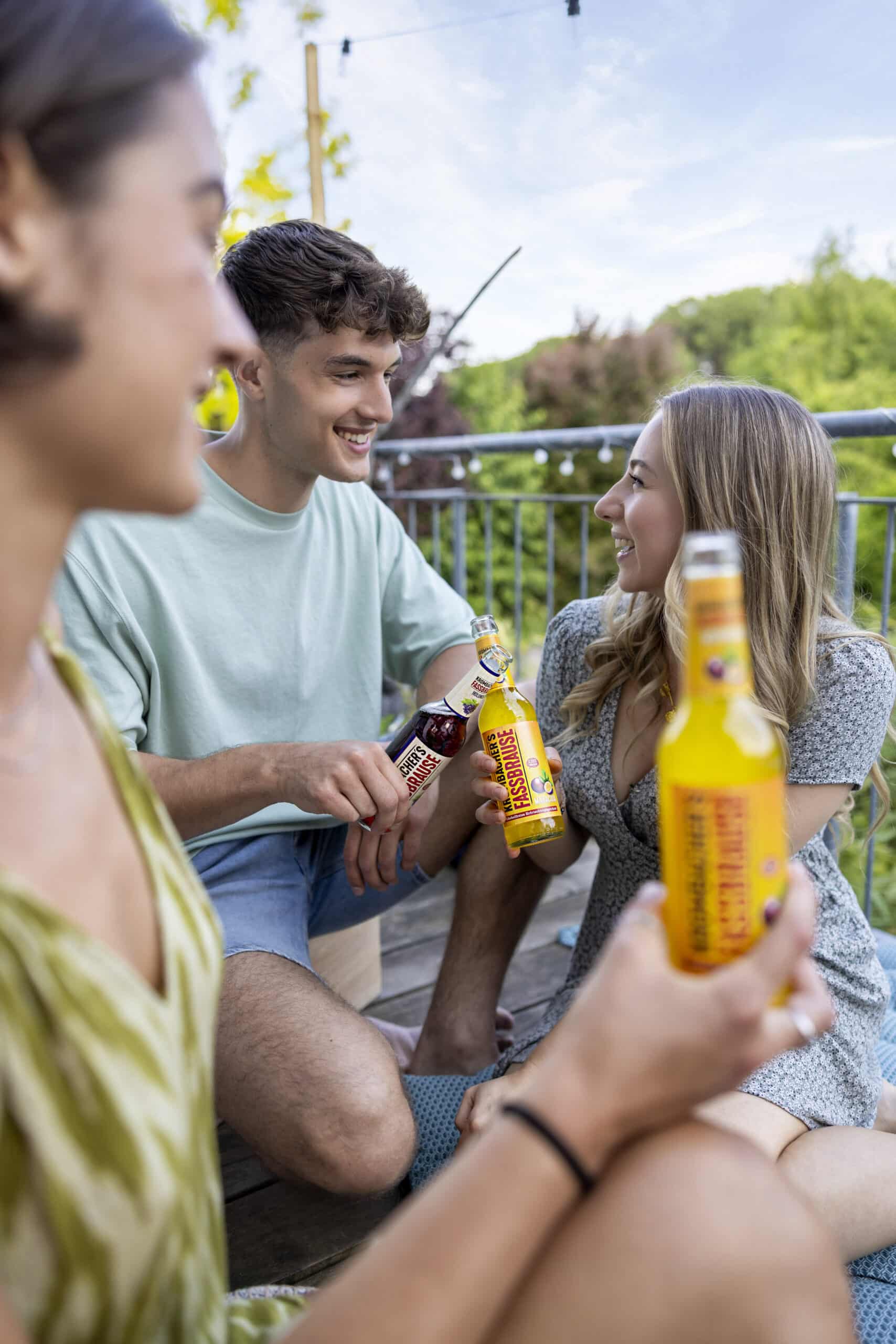 Drei Freunde treffen sich gemütlich im Freien und halten jeweils eine Flasche Limonade in der Hand. Sie sitzen auf einer Holzterrasse mit Grün im Hintergrund, lächeln und unterhalten sich. Der Fokus liegt auf ihren fröhlichen Gesichtern und den Limonadenflaschen. © Fotografie Tomas Rodriguez