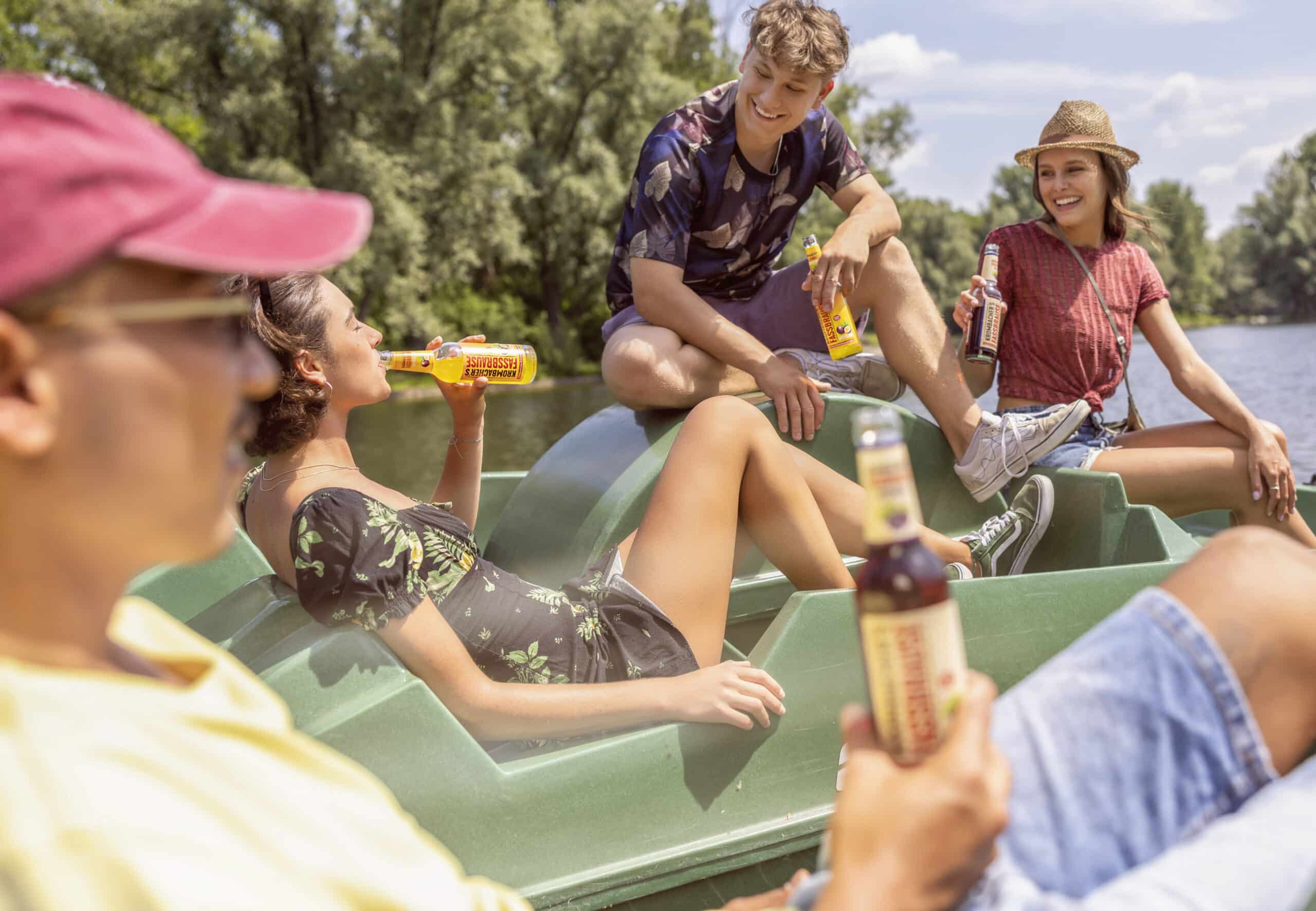 Vier Freunde entspannen an einem sonnigen Tag auf einem Boot. Eine Person in gelbem Hemd und roter Mütze sitzt im Vordergrund, während zwei andere auf dem Deck sitzen. Alle genießen Flaschengetränke vor der malerischen Kulisse von Bäumen und Wasser. © Fotografie Tomas Rodriguez