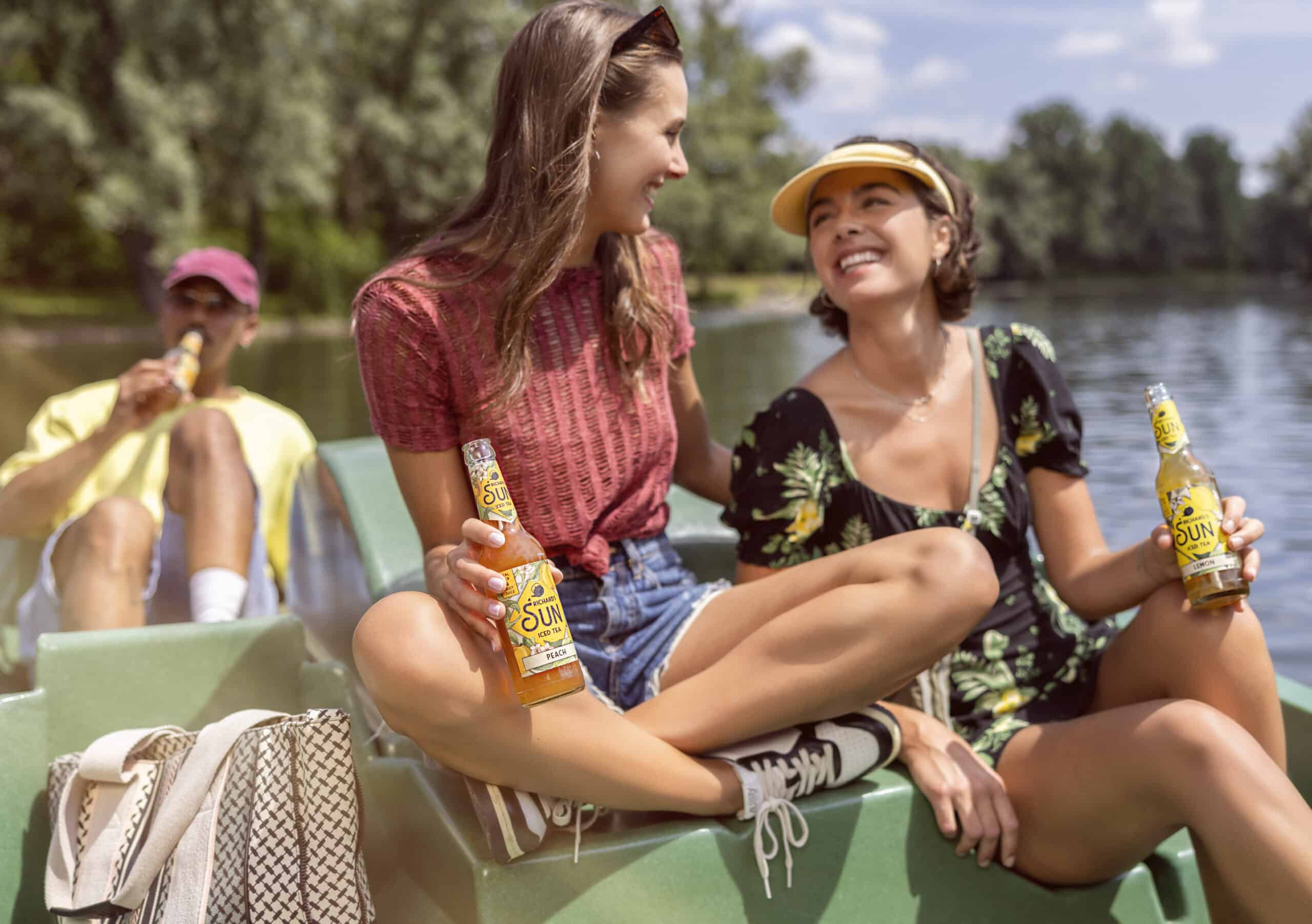 Drei Freundinnen genießen einen sonnigen Tag auf einem Boot. Zwei Frauen sitzen und lächeln und halten Flaschen mit SUN-Getränken in der Hand. Eine trägt ein rotes Top und Shorts, die andere ein geblümtes Kleid und eine gelbe Schirmmütze. Im Hintergrund ist ein Mann in einem gelben Hemd und einer Kappe, ebenfalls mit einem Getränk. © Fotografie Tomas Rodriguez