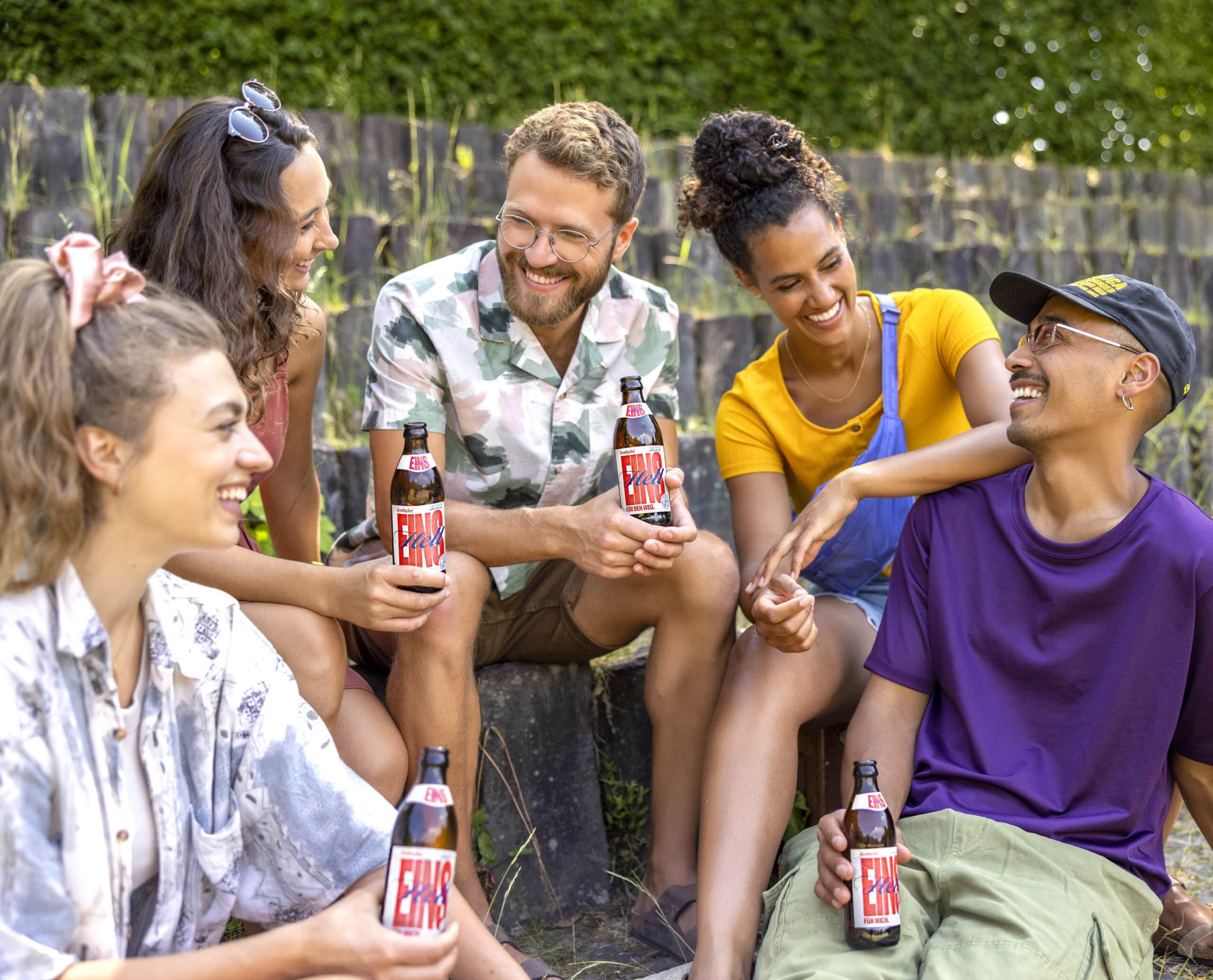 Eine Gruppe von fünf Freunden sitzt auf Steintreppen im Freien, lächelt und lacht gemeinsam, während sie Getränkeflaschen in den Händen halten. Sie sind leger gekleidet und genießen einen sonnigen Tag, umgeben von viel Grün. © Fotografie Tomas Rodriguez