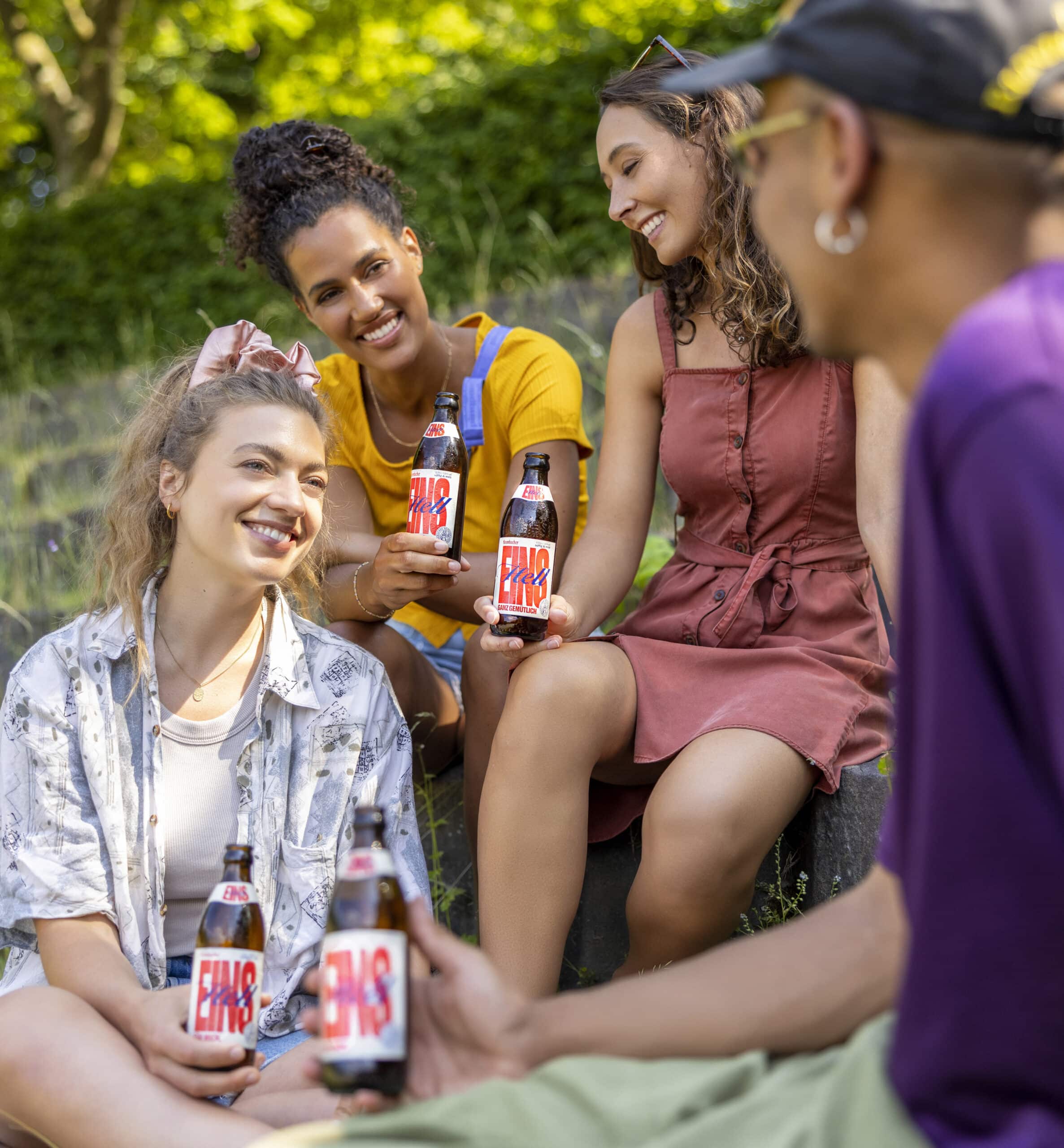 Vier Menschen sitzen an einem sonnigen Tag im Freien, lächeln und halten Flaschen mit einem Getränk mit der Aufschrift „Fass Brause“ in den Händen. Sie scheinen ein lockeres, geselliges Beisammensein in einer parkähnlichen Umgebung mit Gras und Bäumen im Hintergrund zu genießen. © Fotografie Tomas Rodriguez