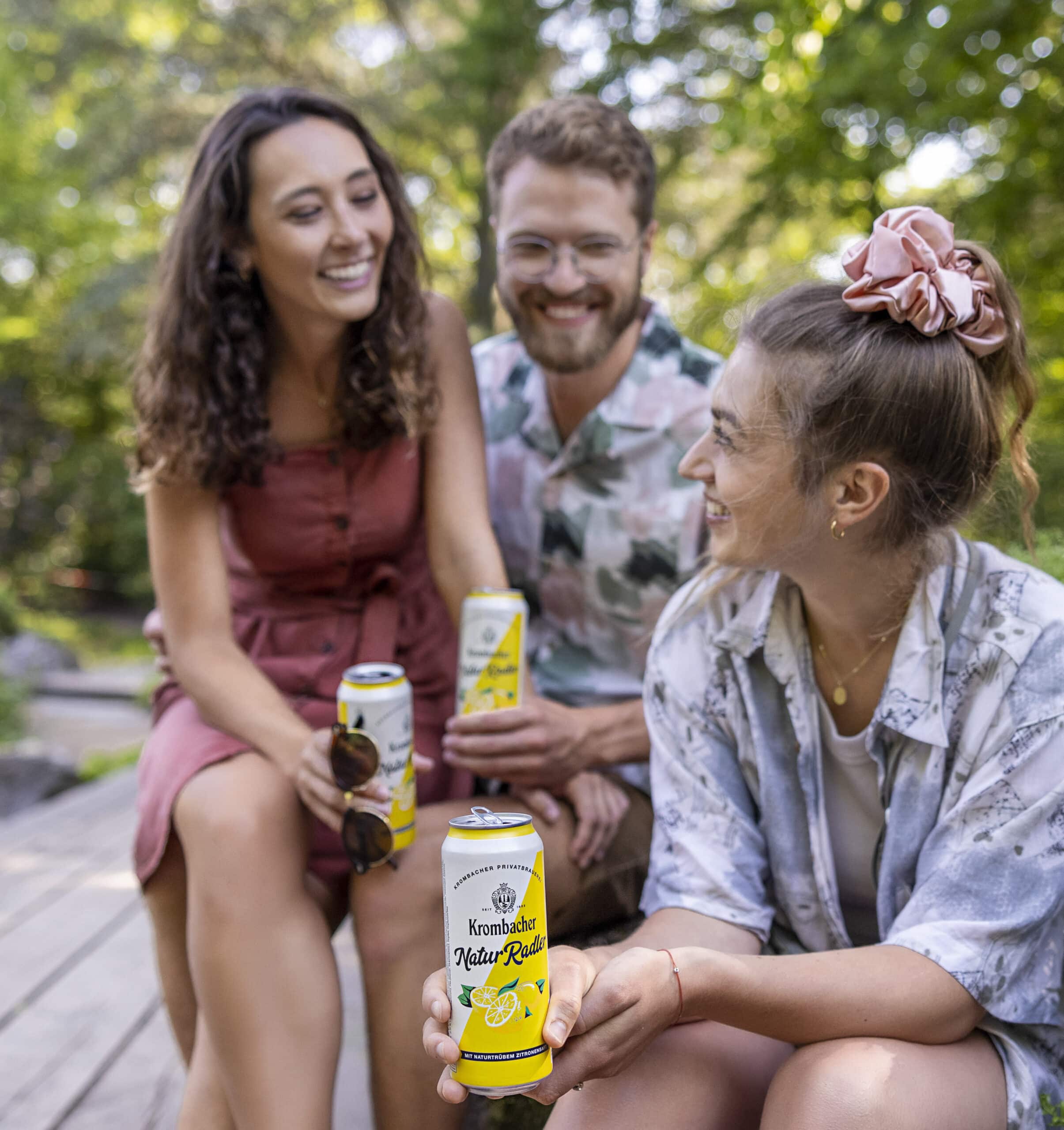 Drei Leute sitzen draußen und genießen Getränke aus der Dose. Sie lächeln alle und scheinen Spaß zu haben. Zwei Frauen, eine mit einem Haargummi im Haar, und ein Mann in der Mitte halten gelb-weiße Dosen mit einem Zitronenmotiv. Im Hintergrund sind Bäume zu sehen. © Fotografie Tomas Rodriguez