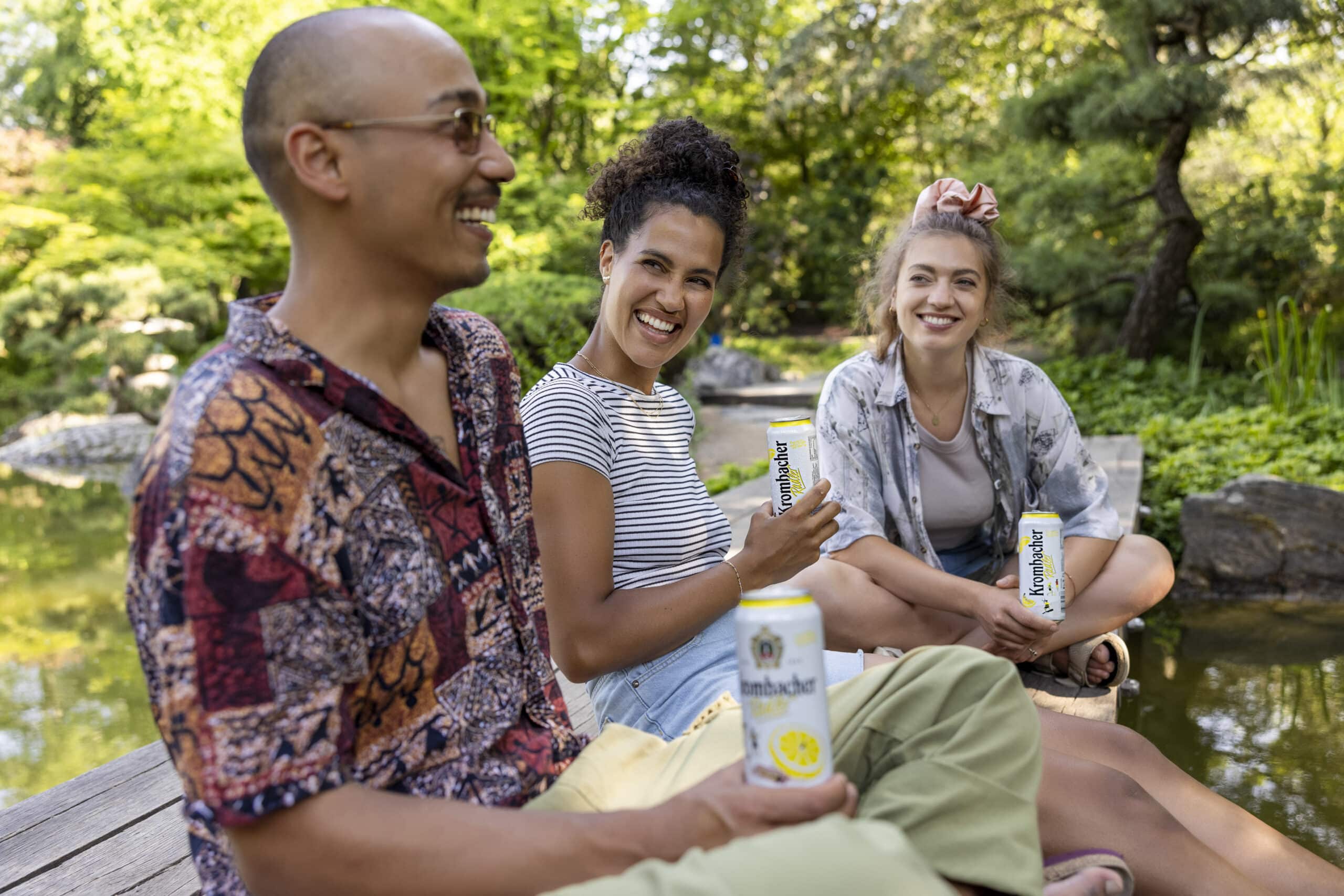 Drei Personen sitzen auf einem Holzdeck neben einem Teich in einem üppigen Garten, lächeln und halten Dosen Krombacher-Bier in den Händen. Die Gruppe scheint einen sonnigen, entspannten Ausflug im Grünen zu genießen. © Fotografie Tomas Rodriguez
